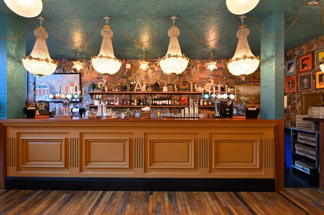 Elegant Cadno bar interior with a long wooden counter, ornate chandeliers, and decorative wallpaper. The back wall displays bottles, glasses, and framed art, while star-shaped lights hang above the bar. The floor is wooden.