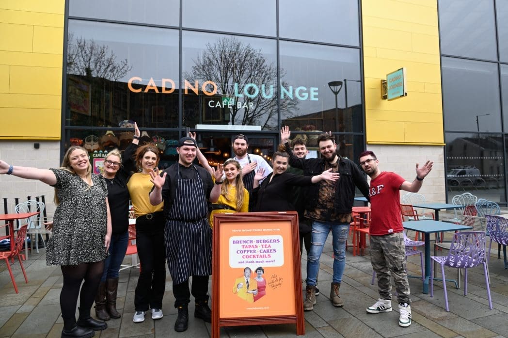 A group of people, some in work uniforms, pose and smile with arms raised outside Cadno Lounge. Colorful Cadno chairs and tables are arranged outdoors, while a signboard advertises food and drinks.