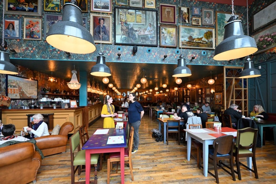 A lively café with eclectic wall art, vintage decor, and hanging lights; people are seated at tables eating and chatting, while two people stand talking in the center of the Cadno-themed room.