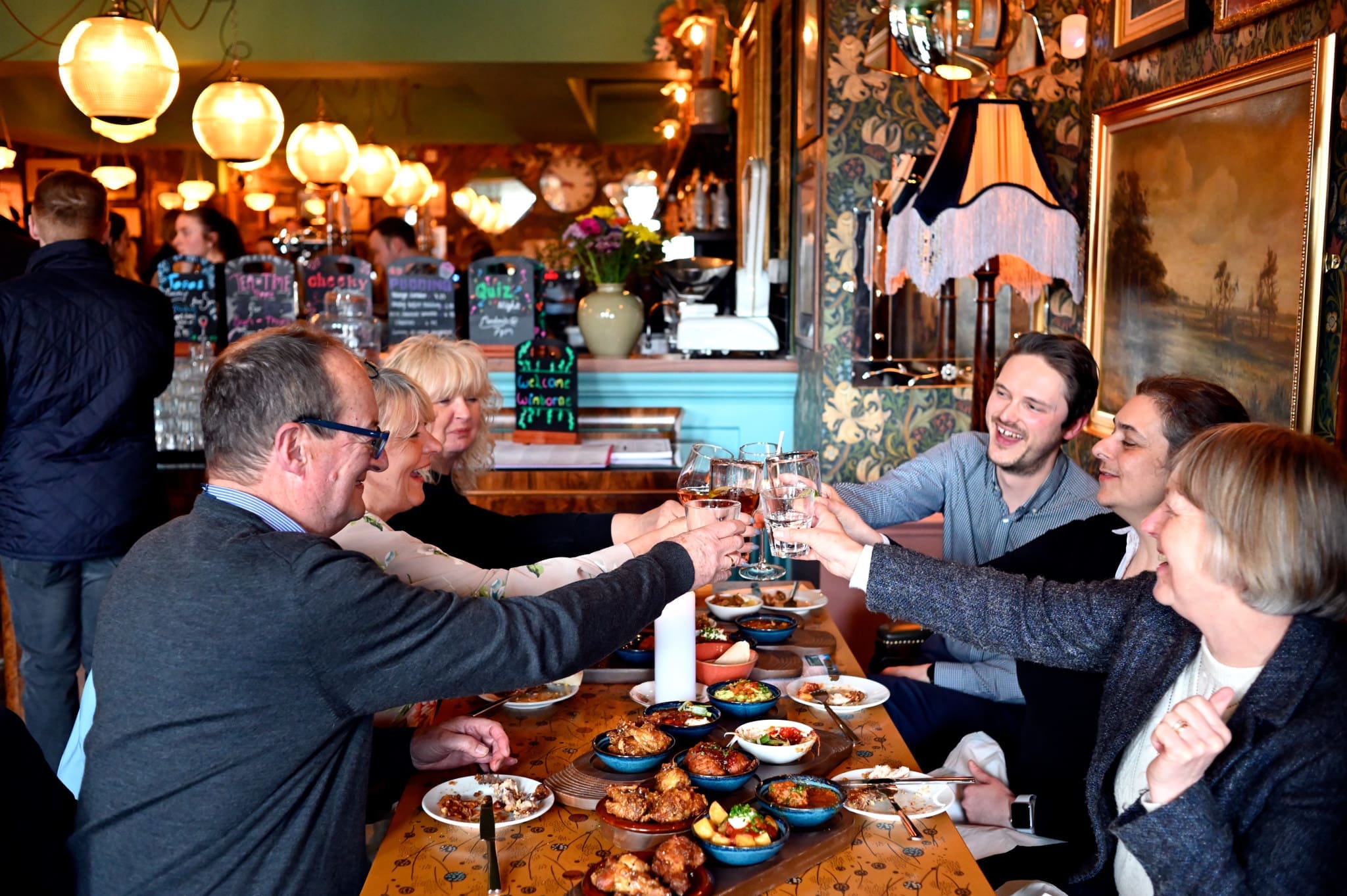 Six people sit around a restaurant table filled with food, raising their glasses in a toast and smiling, while other patrons enjoy the warmly lit Piazzo, beautifully decorated in the background.