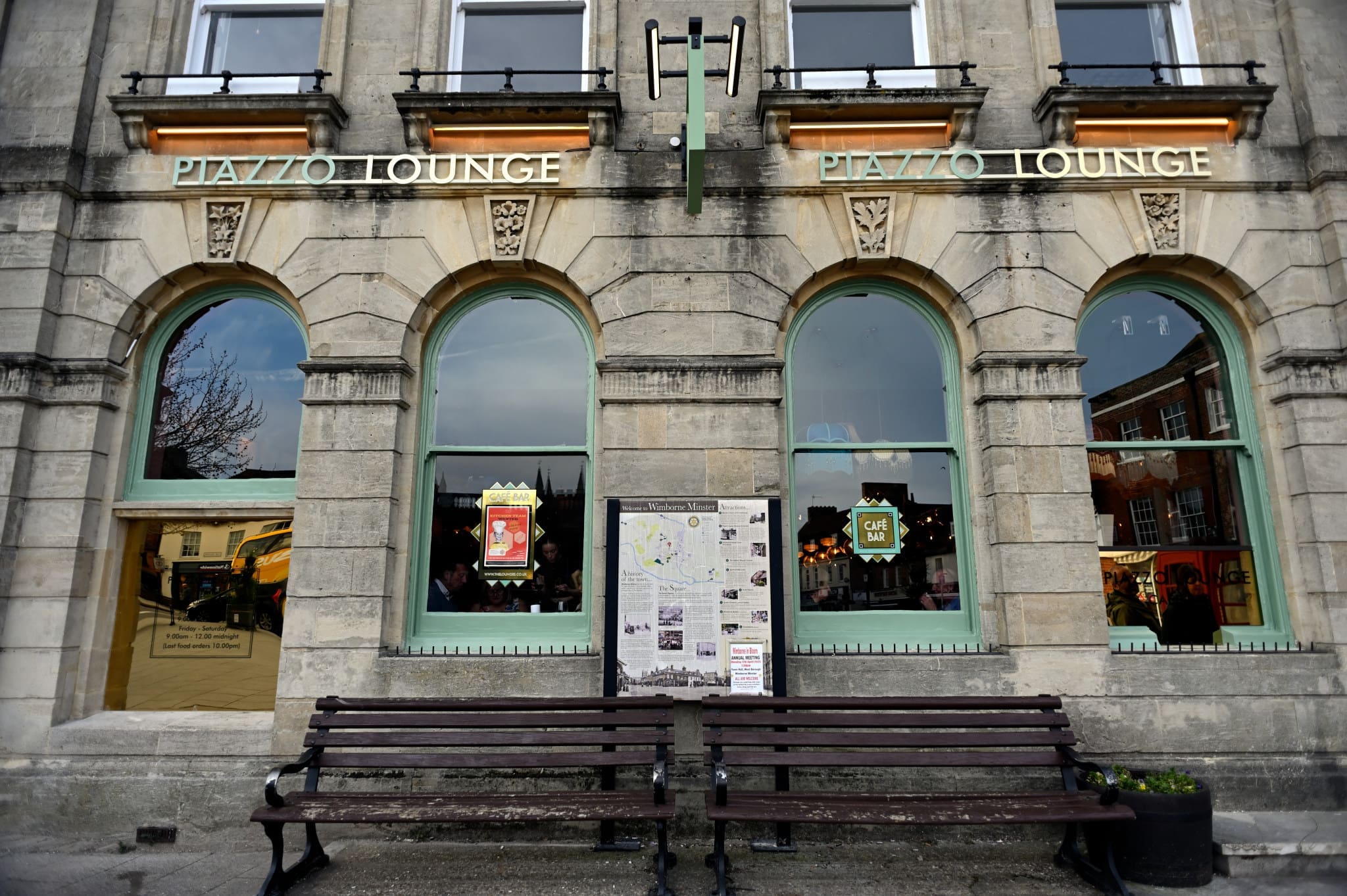 A stone building with arched windows and “PIAZZO LOUNGE” signs above them. Two benches and an informational display board sit in front. Warm lights and Piazzo-inspired decor are visible inside through the windows.