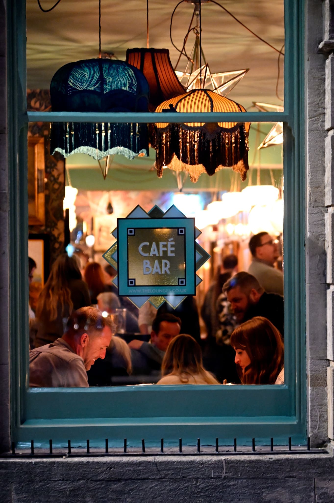 A view through a café bar window shows people sitting and talking inside. Warm lighting, decorative lamps, and a sign reading "CAFÉ BAR" create a cozy atmosphere reminiscent of a Piazzo gathering.