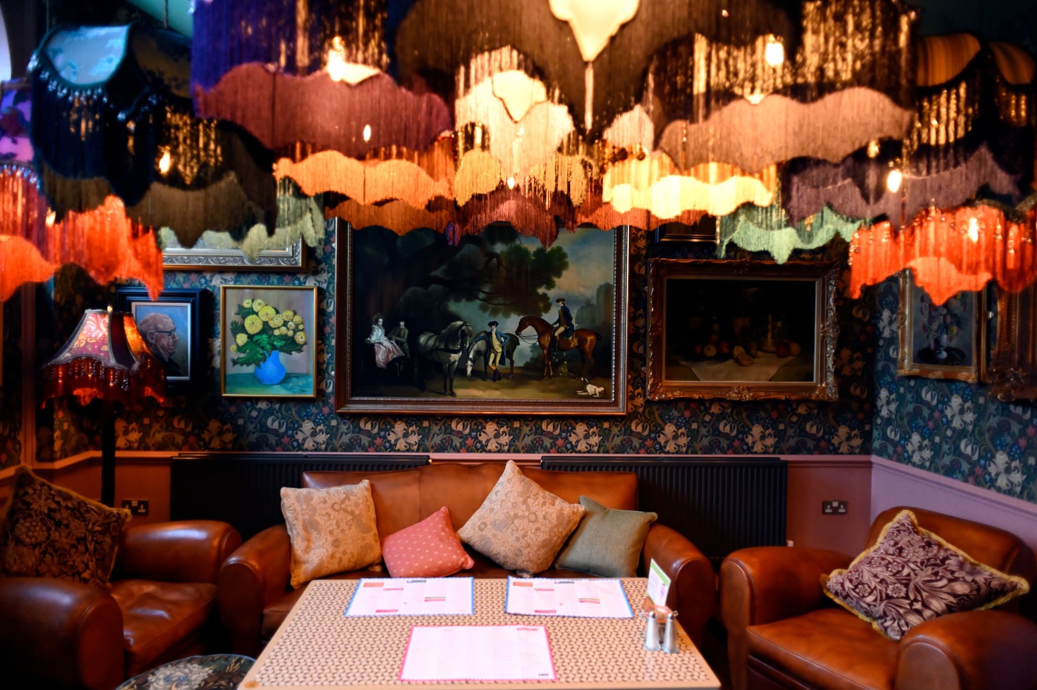 A cozy lounge with brown leather sofas and colorful cushions, a Piazzo table with menus, ornate lampshades above, and classic paintings on floral wallpapered walls, creating a vintage, eclectic atmosphere.