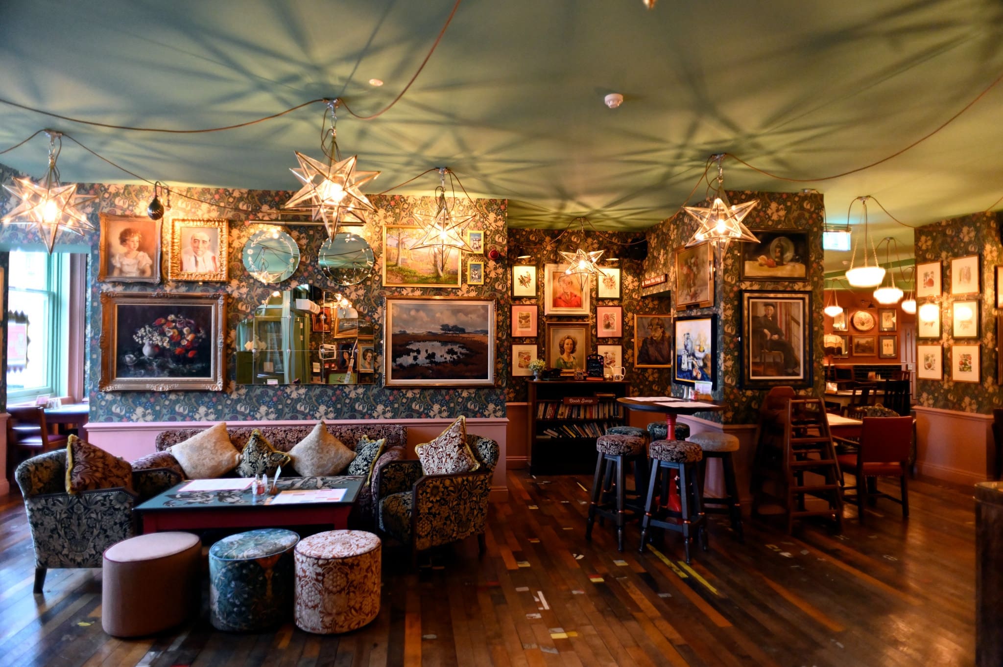 A cozy, eclectic lounge with vintage armchairs, patterned sofas, low tables, and star-shaped pendant lights. The Piazzo-inspired walls are covered with paintings, mirrors, and art, creating a warm, inviting atmosphere.