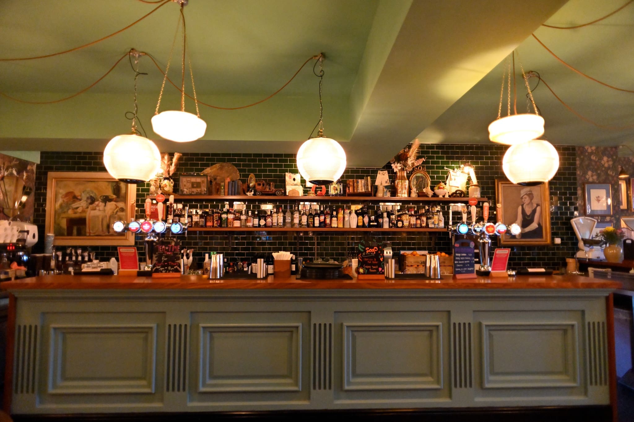 A cozy Piazzo bar with a green tiled wall, shelves filled with bottles, and three round hanging lights above a wooden counter. Framed art and decorative items add to the warm, inviting atmosphere.