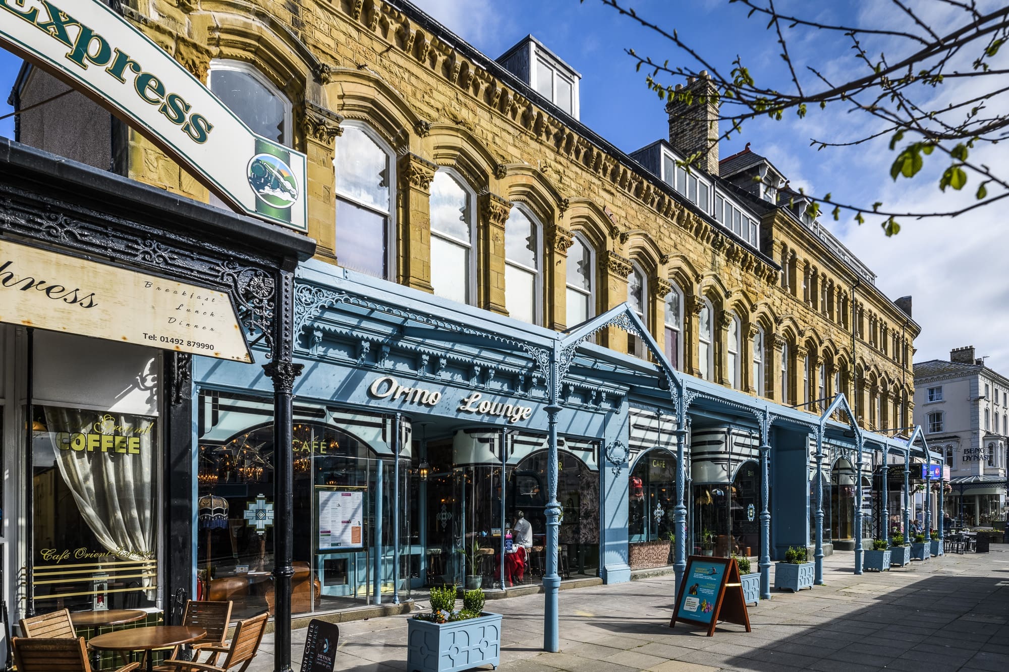 A row of historic stone and blue-painted buildings, including Oppio and "Orno Lounge," houses shops and cafes along a sunny street, where people relax at tables outside under a clear blue sky.