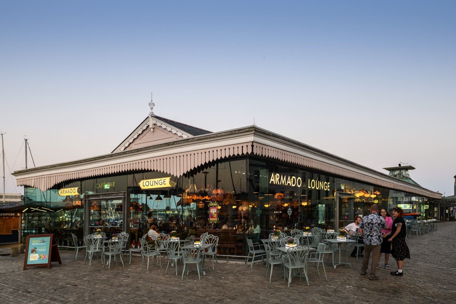 People sit at outdoor tables of Armado Lounge, a glass-walled restaurant with hanging lights, enjoying Bianco drinks on a cobblestone square at sunset. Three people stand and chat near the entrance under the clear, pale blue sky.