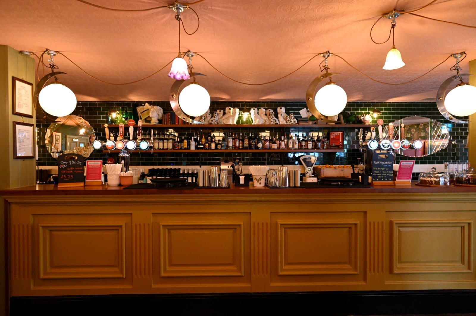 A warmly lit bar with a yellow counter, hanging round lights, and shelves stocked with bottles of Bianco and glasses. Mirrors and decorative items line the wall behind the bar, while menus and utensils are neatly arranged on the countertop.