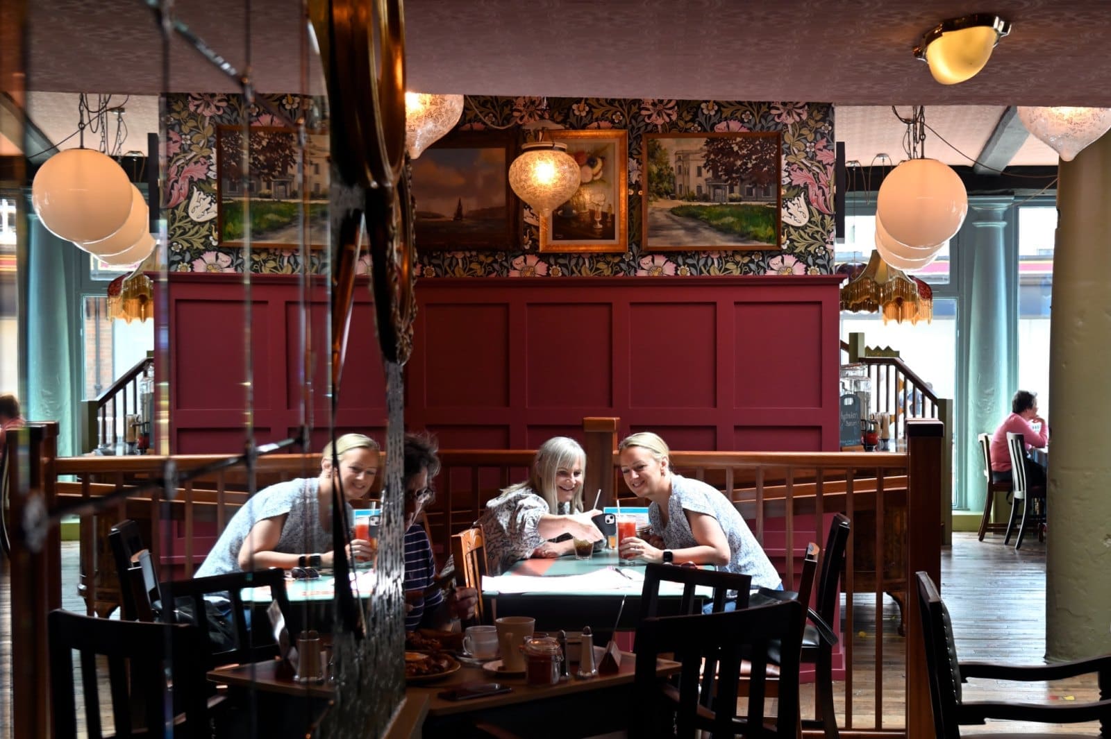 Three people sit together at a table in Bianco, a cozy restaurant with colorful wallpaper, wooden furnishings, and warm lighting from decorative lamps. They smile and look at a phone, enjoying the inviting atmosphere.