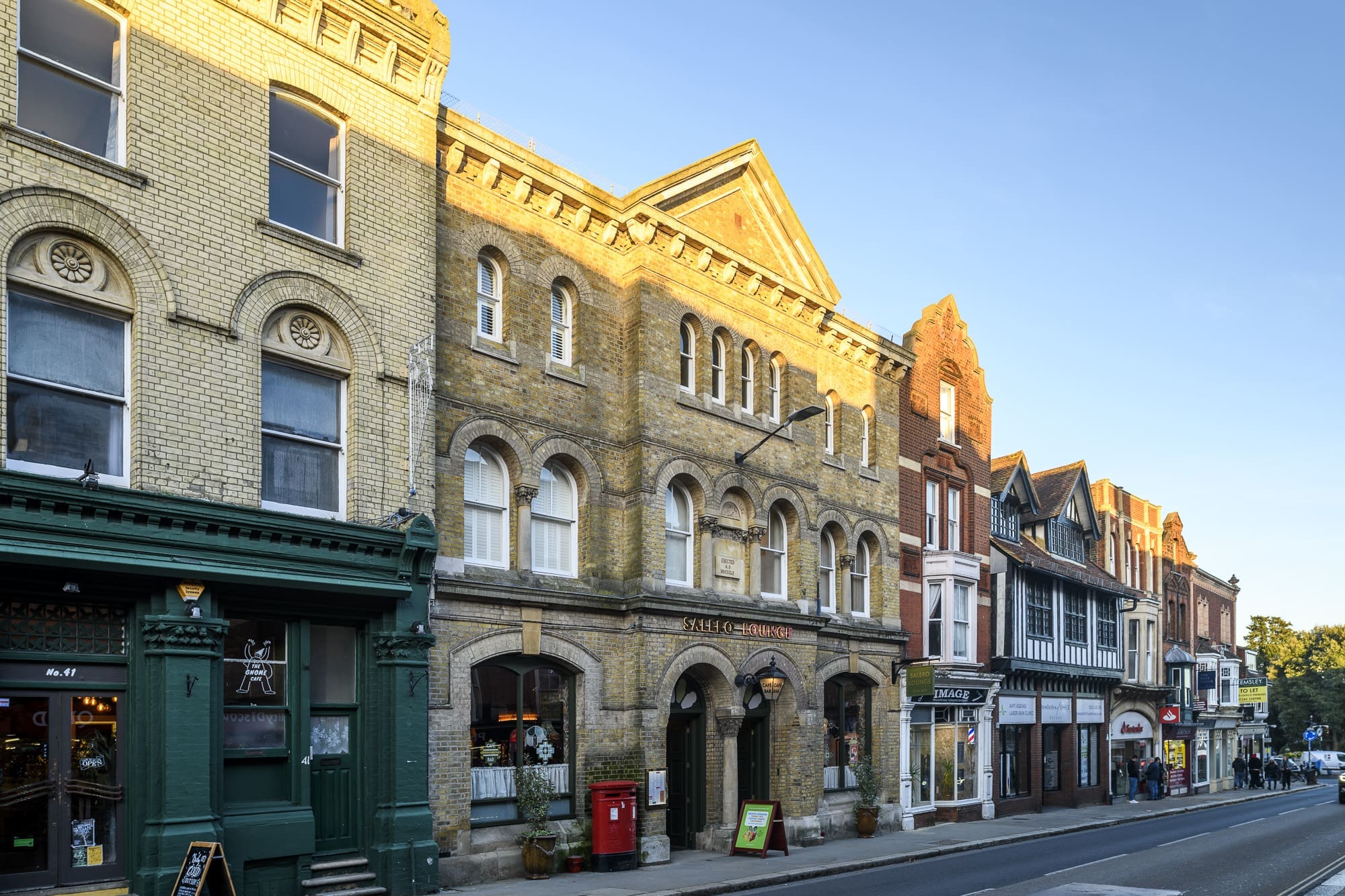 A row of historic, multi-story brick buildings lines a sunny street, with shops, restaurants like Salero, and a post box visible at street level under a clear blue sky.