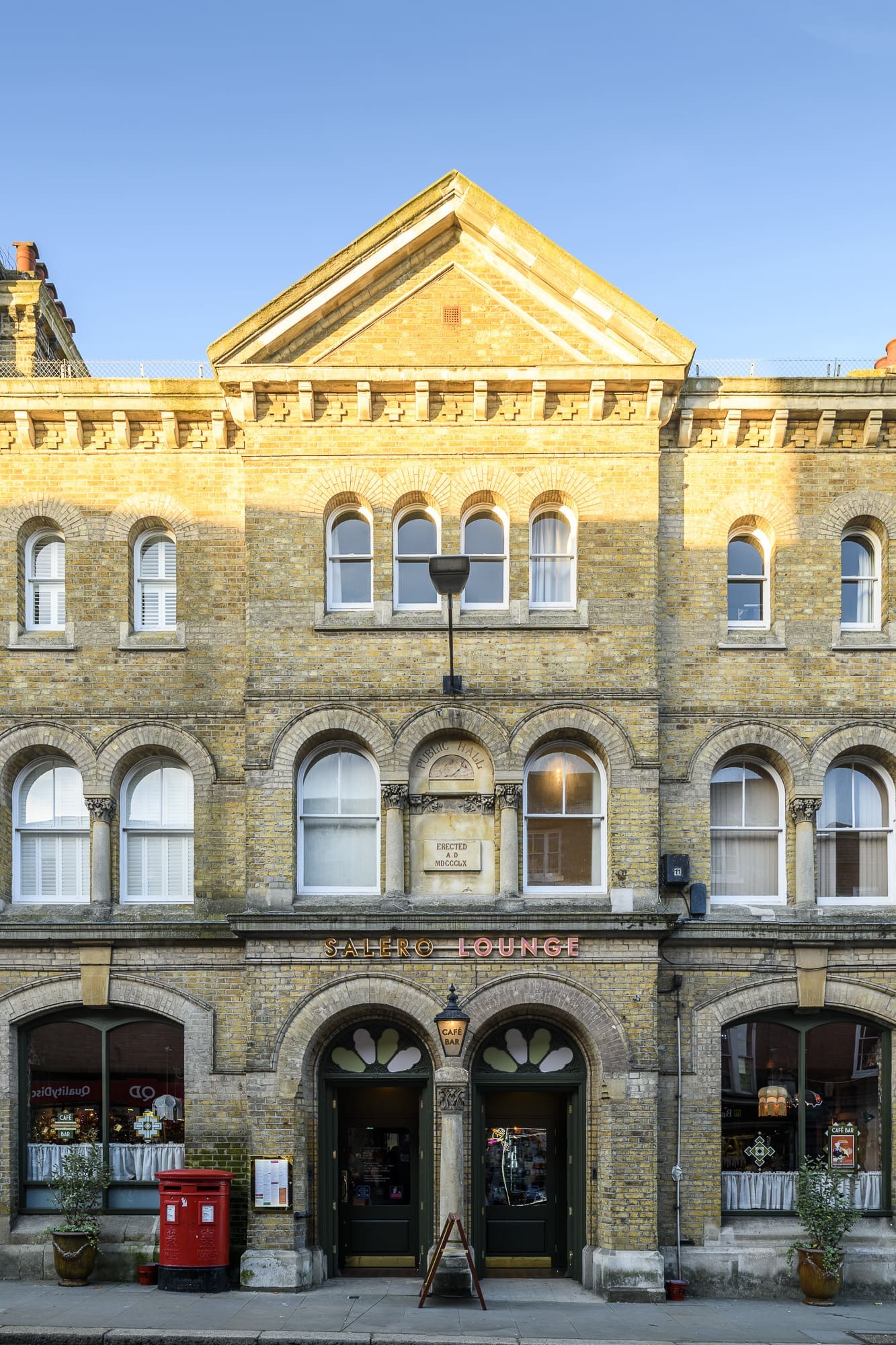 A three-story yellow-brick building with arched windows and a central pediment. The ground floor has two arched doorways marked “Lounge” and shops, including Salero, with a red post box and potted plants on the sidewalk.
