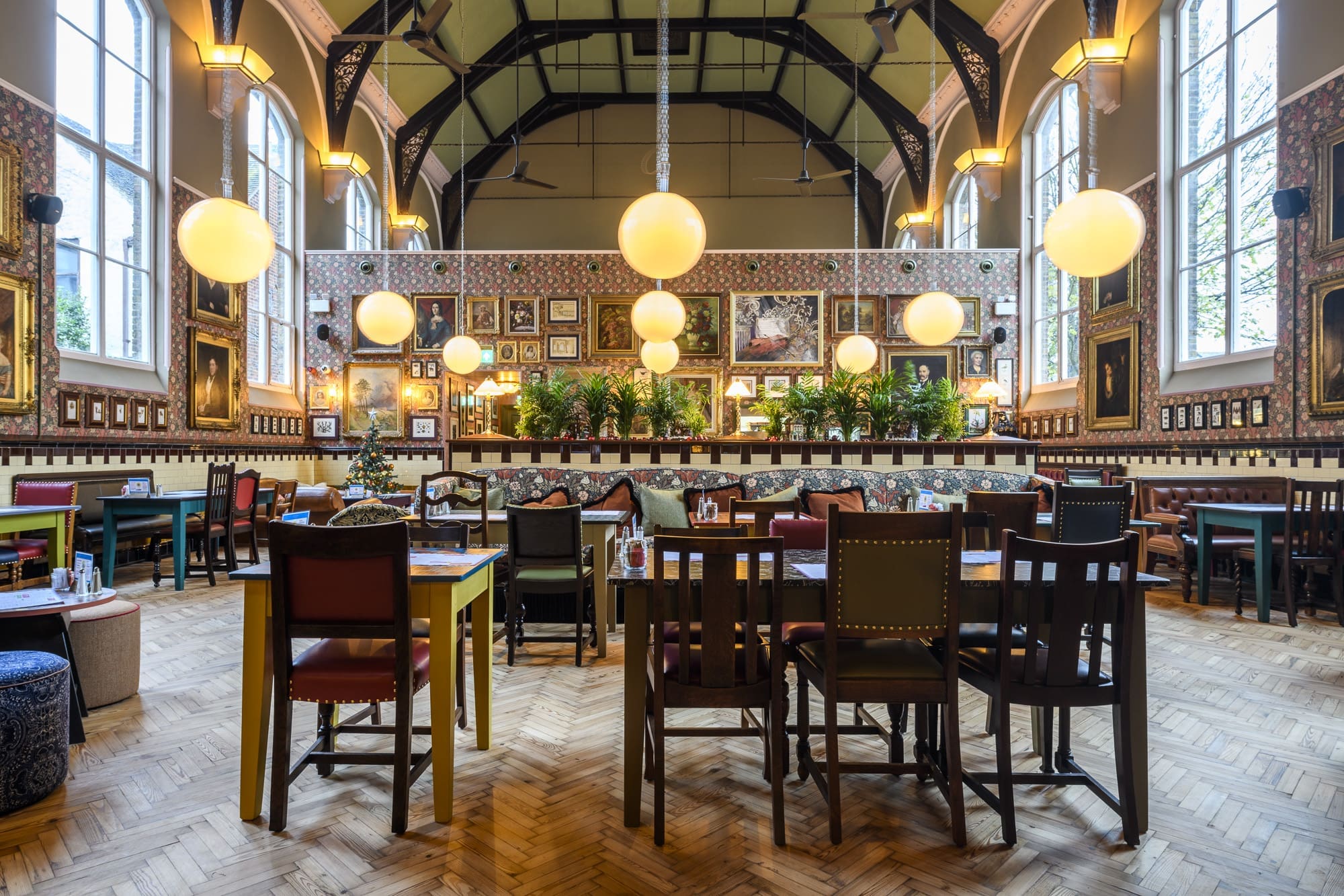 A spacious, stylish restaurant, Salero features high ceilings, large arched windows, globe pendant lights, eclectic wall art, lush greenery, and a mix of colorful tables and chairs arranged neatly on a parquet wood floor.