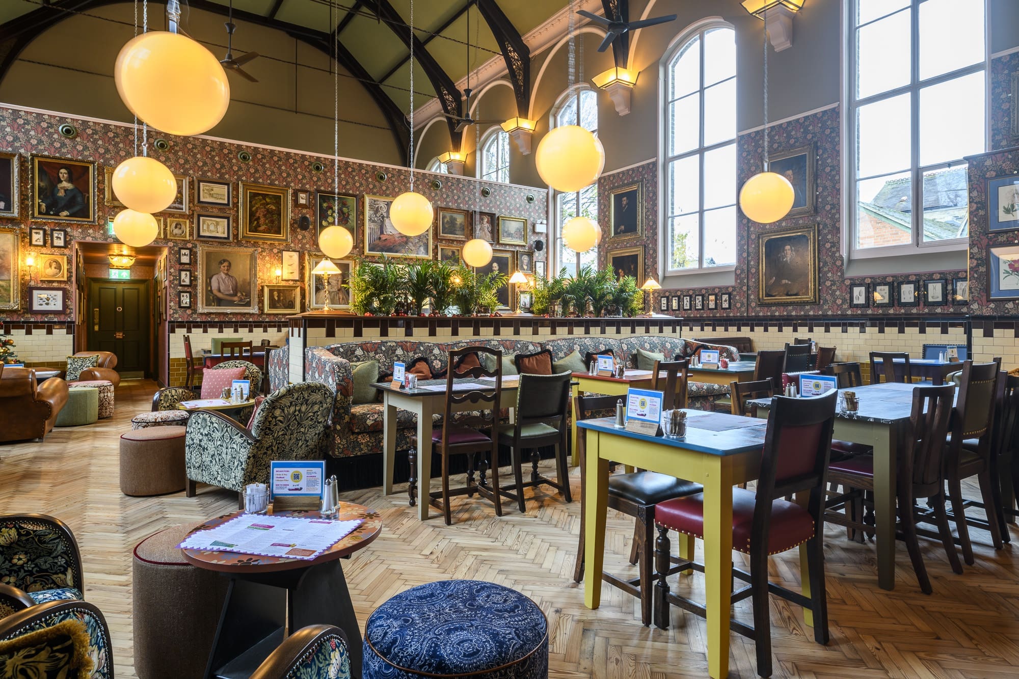 A brightly lit, eclectic restaurant, Salero features hanging globe lights, mismatched chairs and tables, large windows, patterned wallpaper, lush indoor plants, and framed artwork covering the walls.