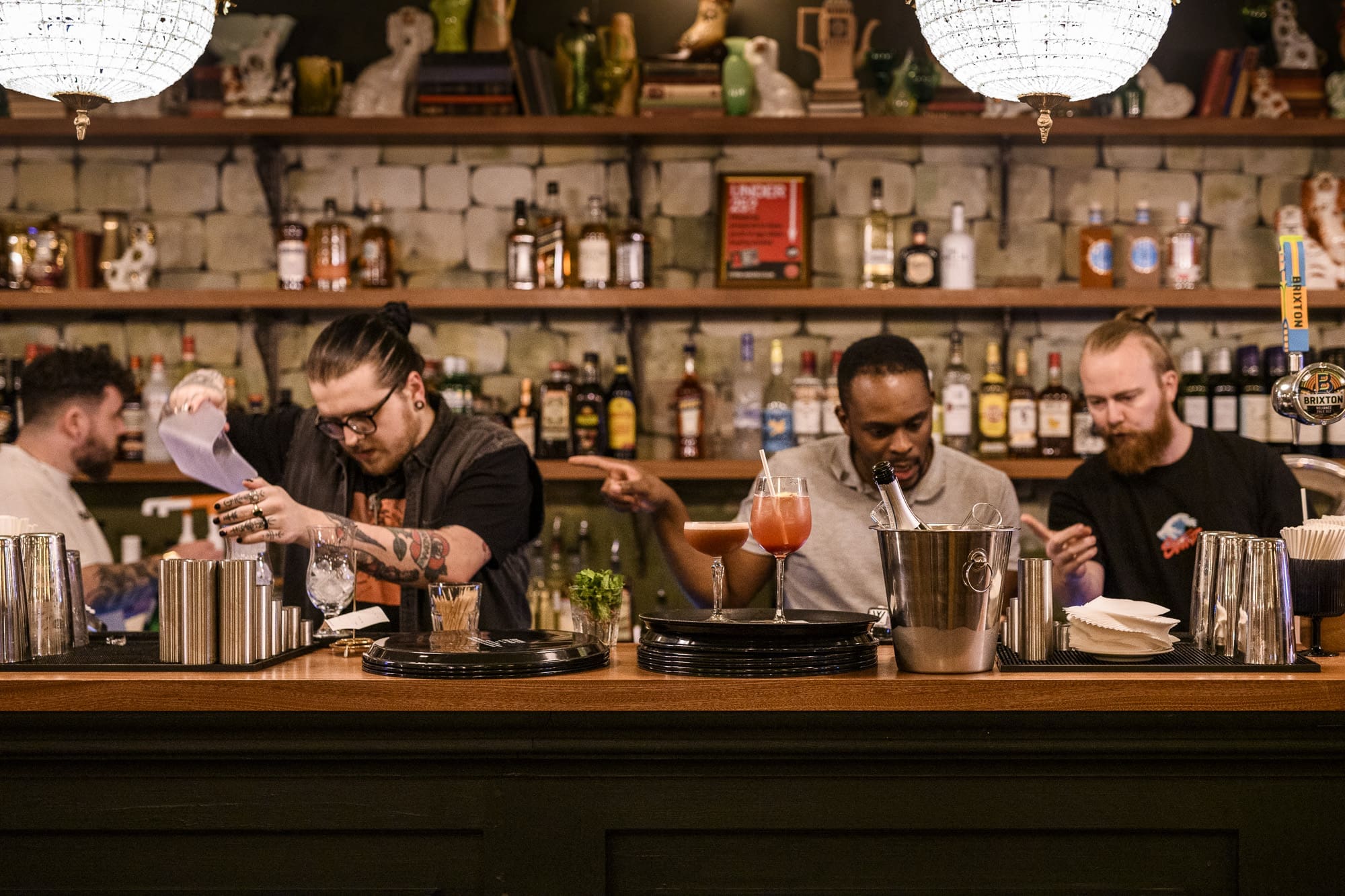 Four bartenders stand behind a bar preparing drinks. Shelves stocked with bottles line the muro behind them. Various bar tools and glasses are on the counter, and one bartender pours a drink while others focus on their tasks.