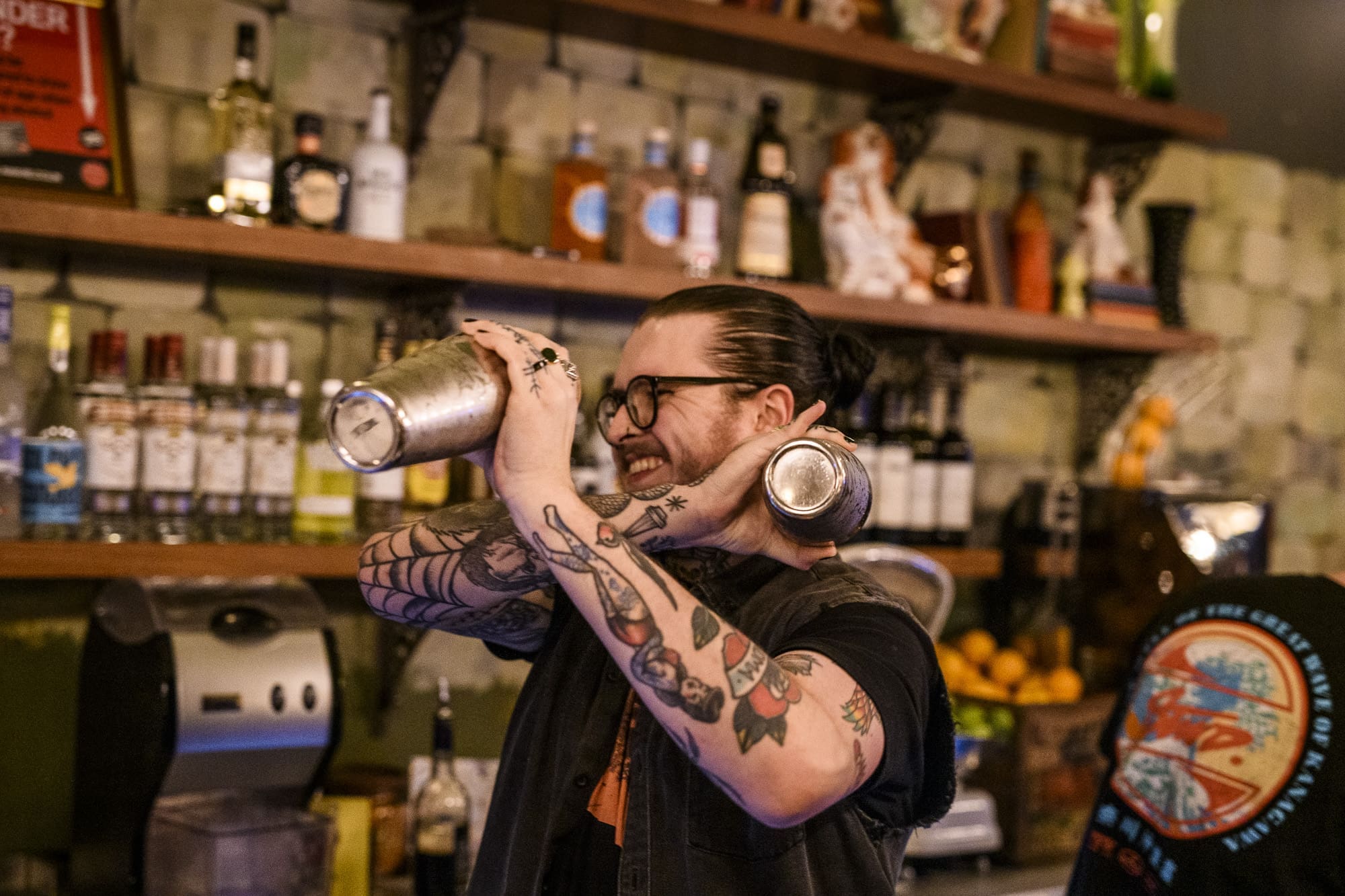A smiling tattooed bartender with glasses shakes a cocktail shaker behind a bar lined with bottles and muro-style decorated shelves.
