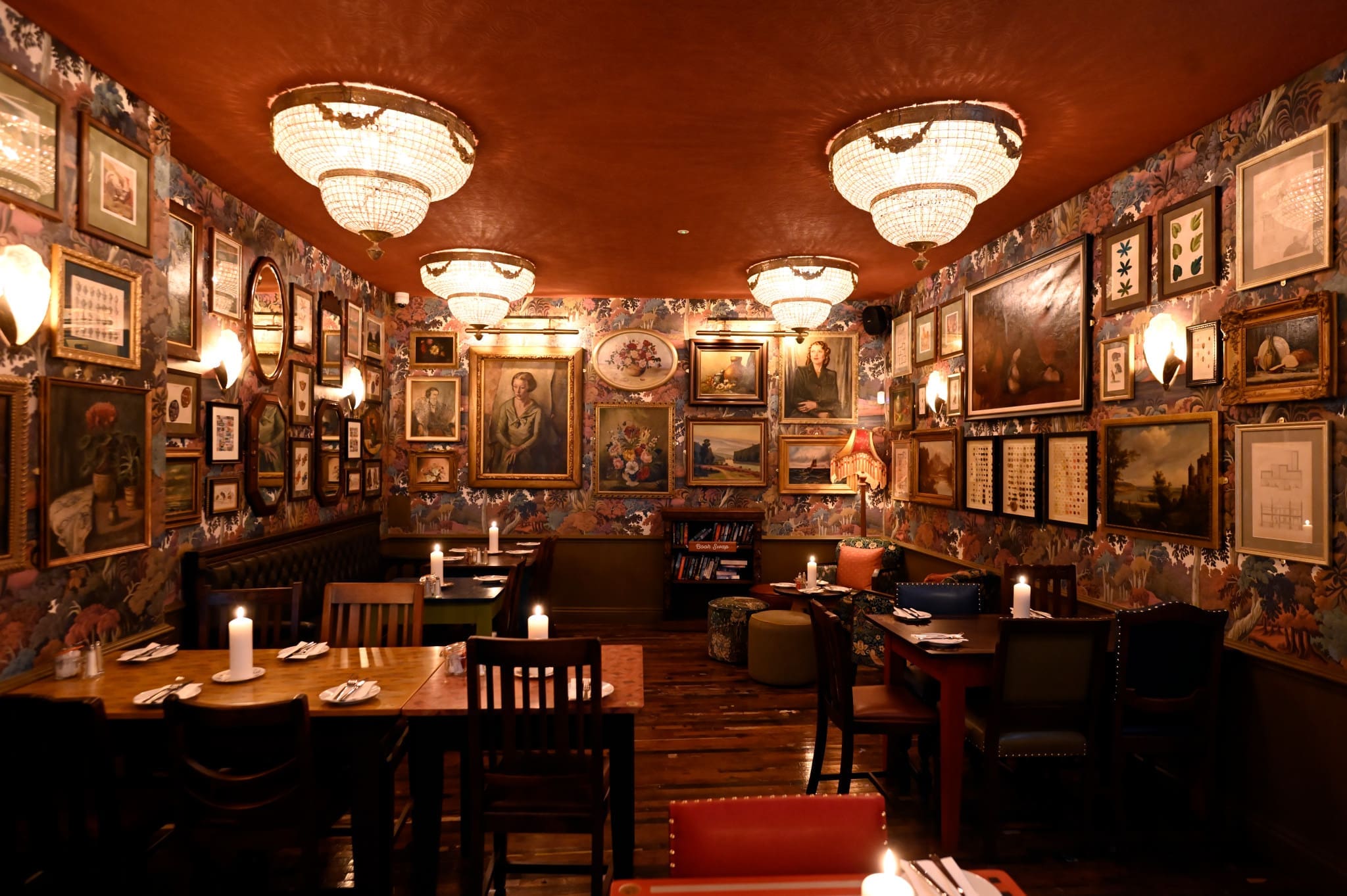A cozy, warmly-lit restaurant near Pontio, with chandeliers, numerous framed paintings and photos covering the floral wallpapered walls, wooden tables set with napkins and candles, and a polished wooden floor.
