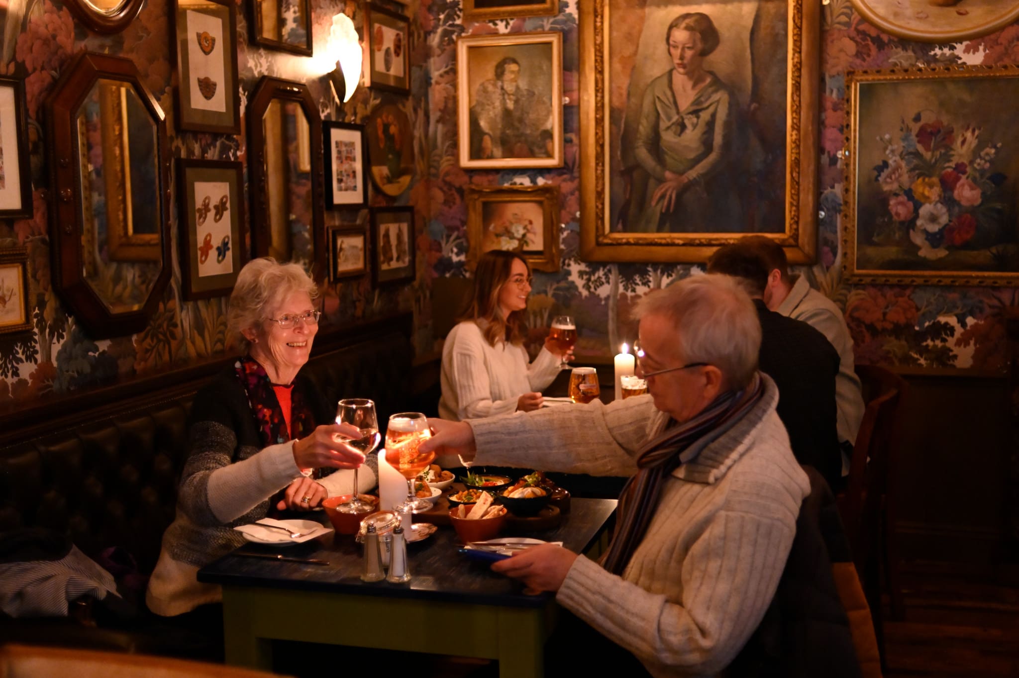 Two older adults clink glasses while dining at Pontio, a cozy, dimly-lit restaurant adorned with vintage paintings and mirrors. Other patrons savor their meals at nearby tables, adding to the warm and inviting atmosphere.