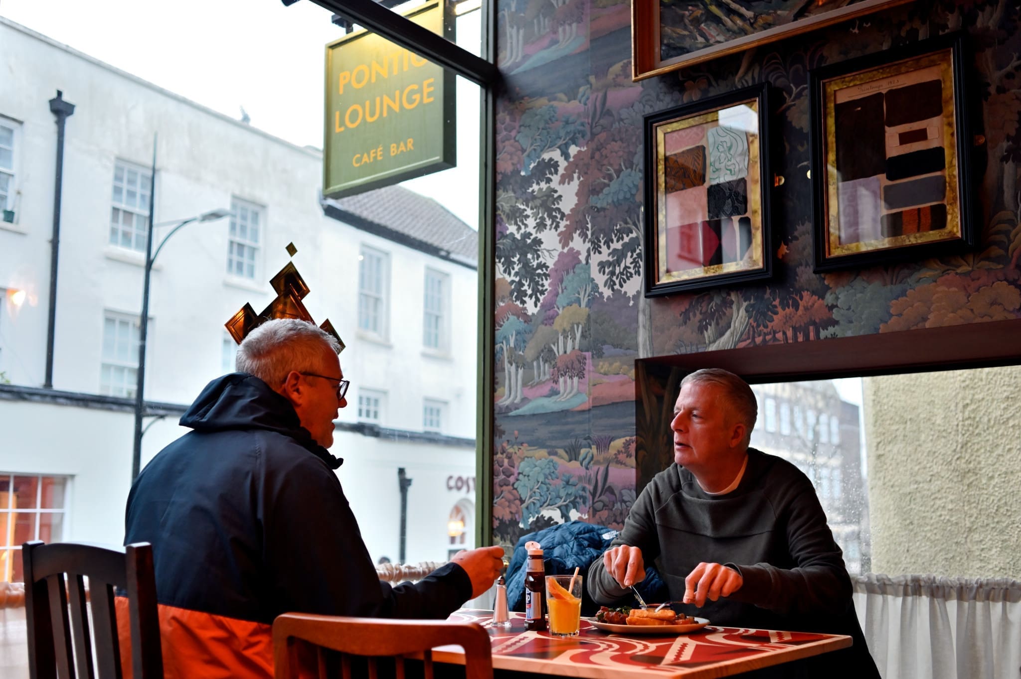 Two men sit across from each other at a table in Pontio café, eating and conversing. One wears glasses and a black jacket; the other has gray hair and a green sweater. Large windows and framed art complete the cozy setting.