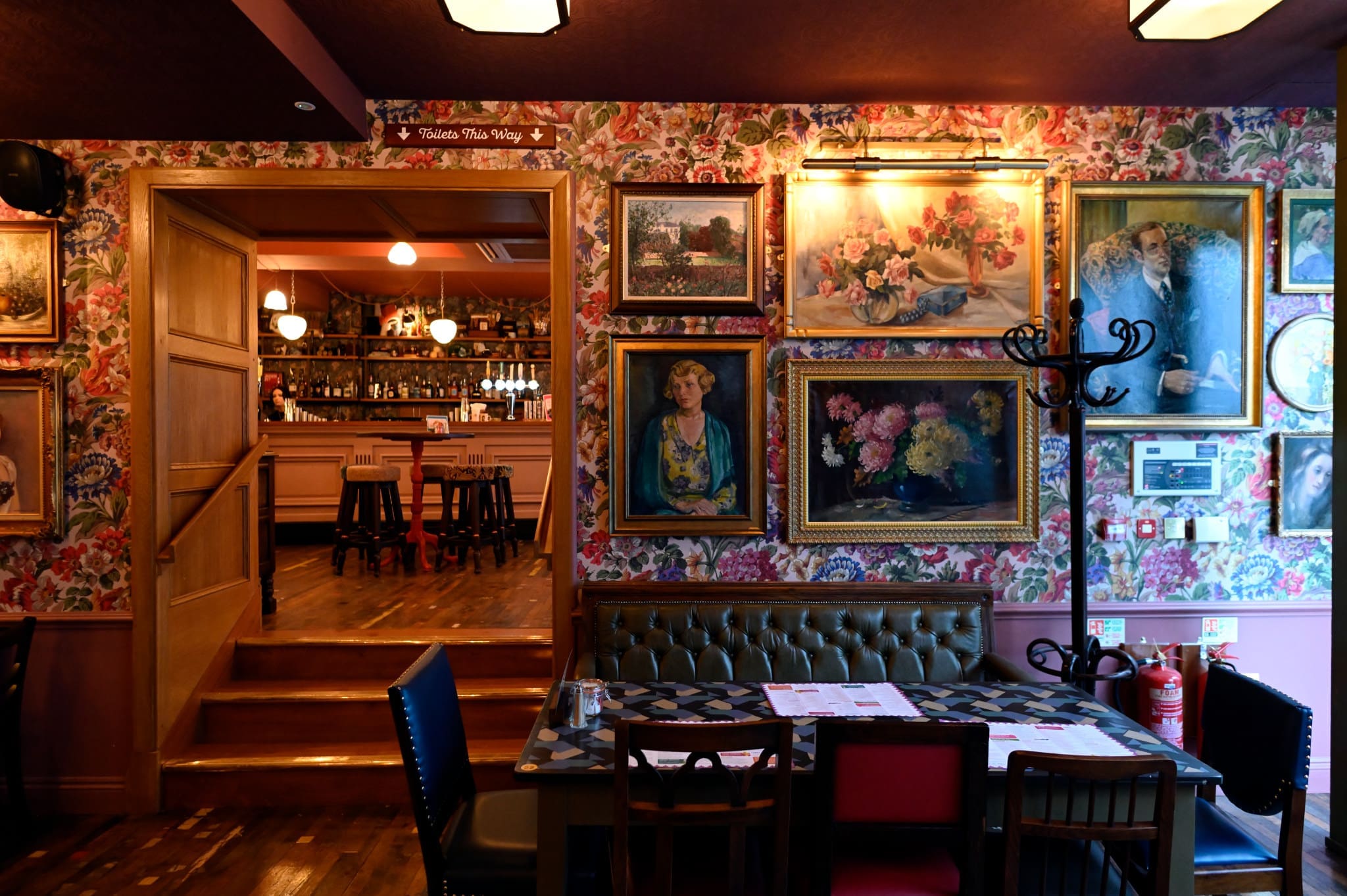 A cozy restaurant interior with floral wallpaper, vintage paintings, a green cushioned booth, mixed chairs around a table with menus, and a bar area visible up a short flight of steps in the stylish pontio district.