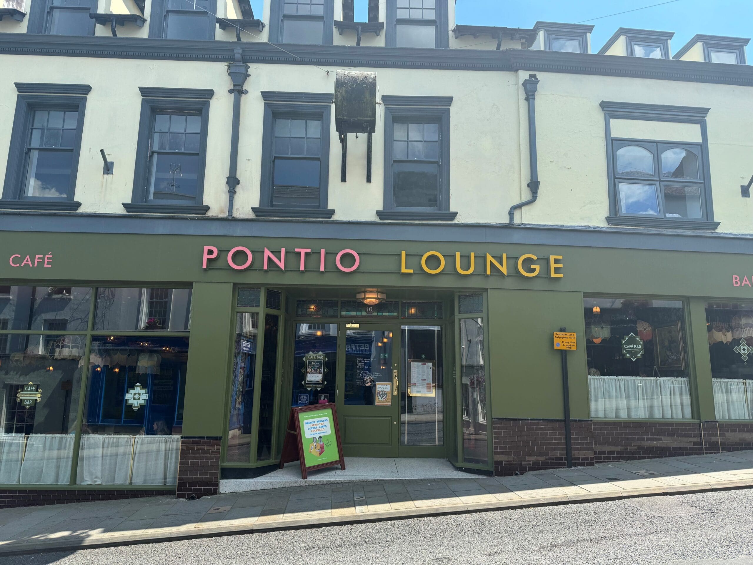 Front view of Pontio Lounge, a cozy café and bar with olive green and brown exterior, large windows, colorful pontio signage, and a sandwich board set on the sunny sidewalk.