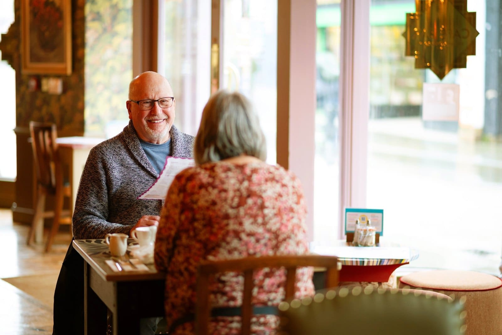 An older man and woman sit across from each other at a cafe table, smiling and talking over coffee. Sunlight streams in through large windows as they savor a glass of Barolo, creating a warm and inviting atmosphere.