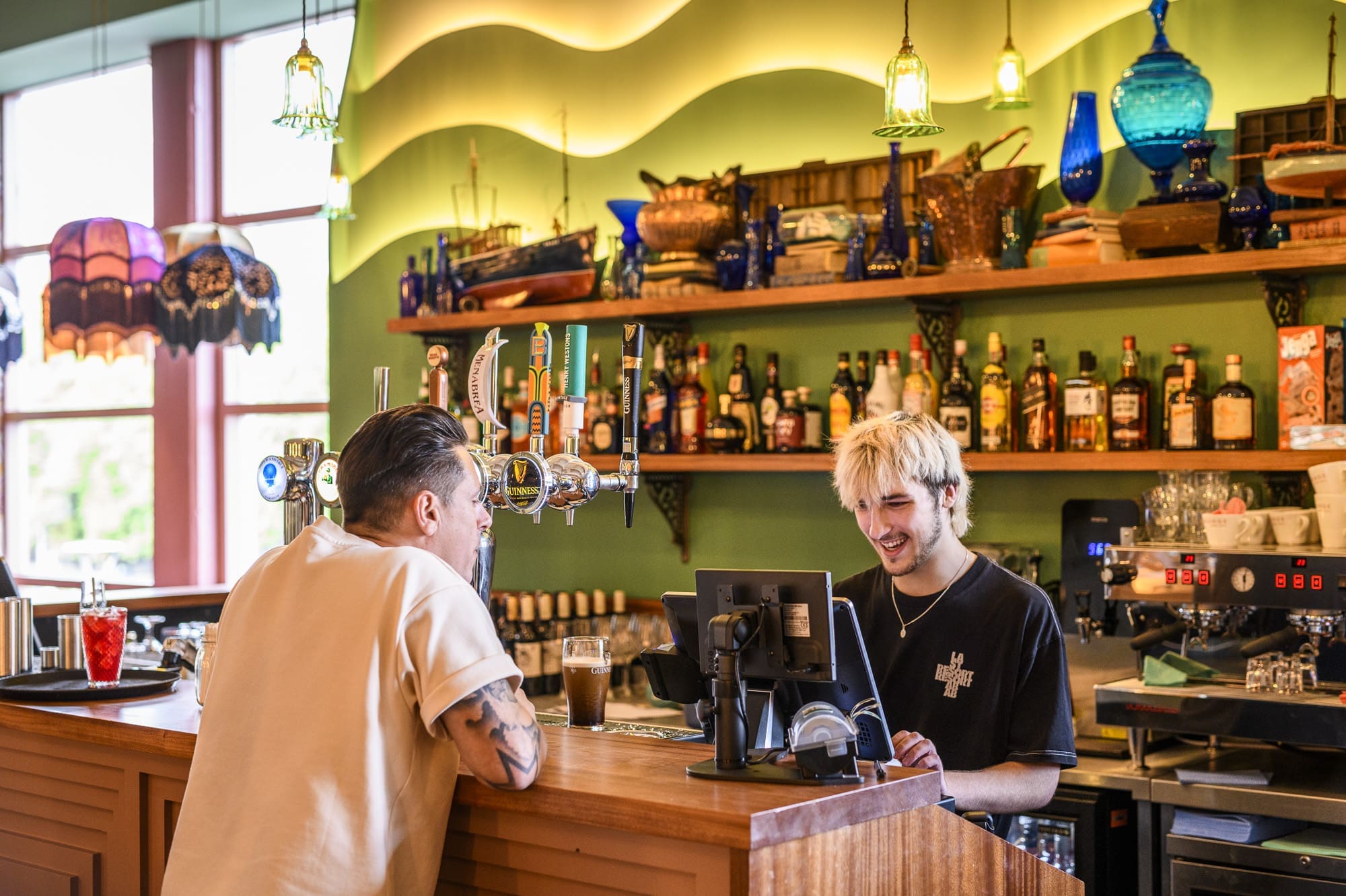 A bartender with bleached hair smiles while taking an order from a customer at Ritorno, a colorful, well-lit bar with bottles, glassware, and decorative items on shelves behind them.