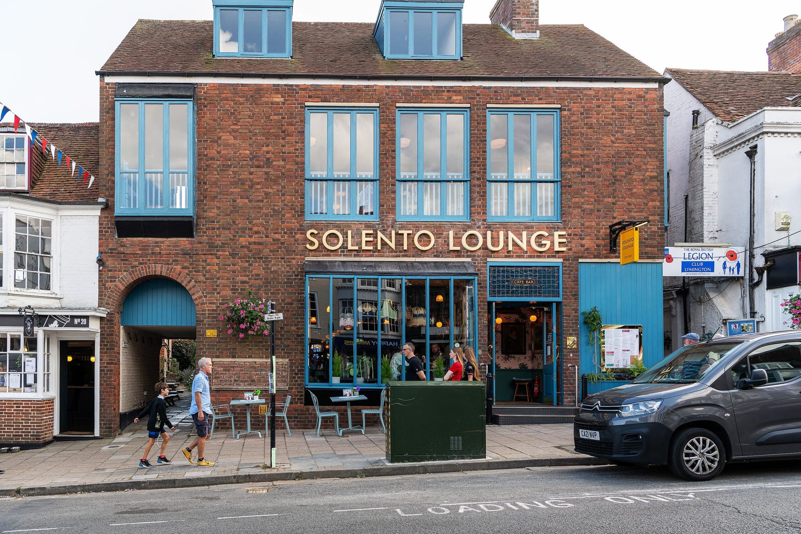 Street view of Solento Lounge, a modern café-bar in a brick building with large blue windows. People stroll past Solento’s outdoor tables, while a car is parked nearby and colorful bunting decorates the left side.