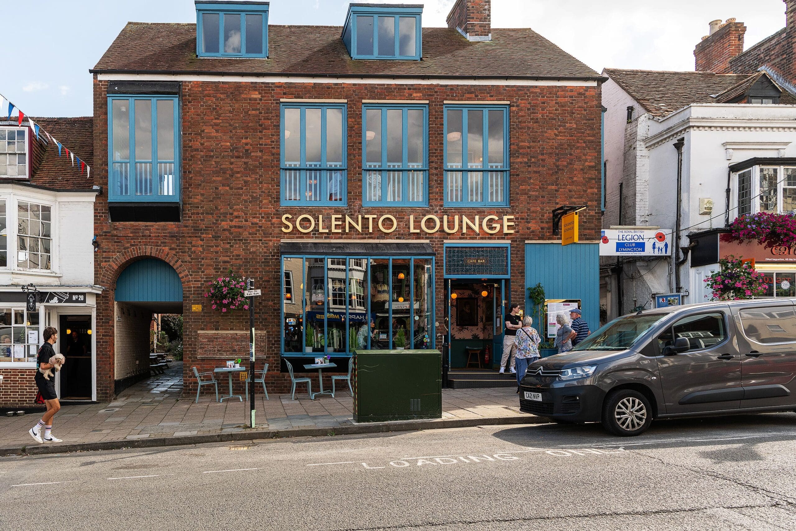 A brick building with teal trim houses Solento Lounge, a vibrant café bar with outdoor seating. People stroll by Solento as a grey van is parked out front, and the lively street and neighboring shops add to the scene.