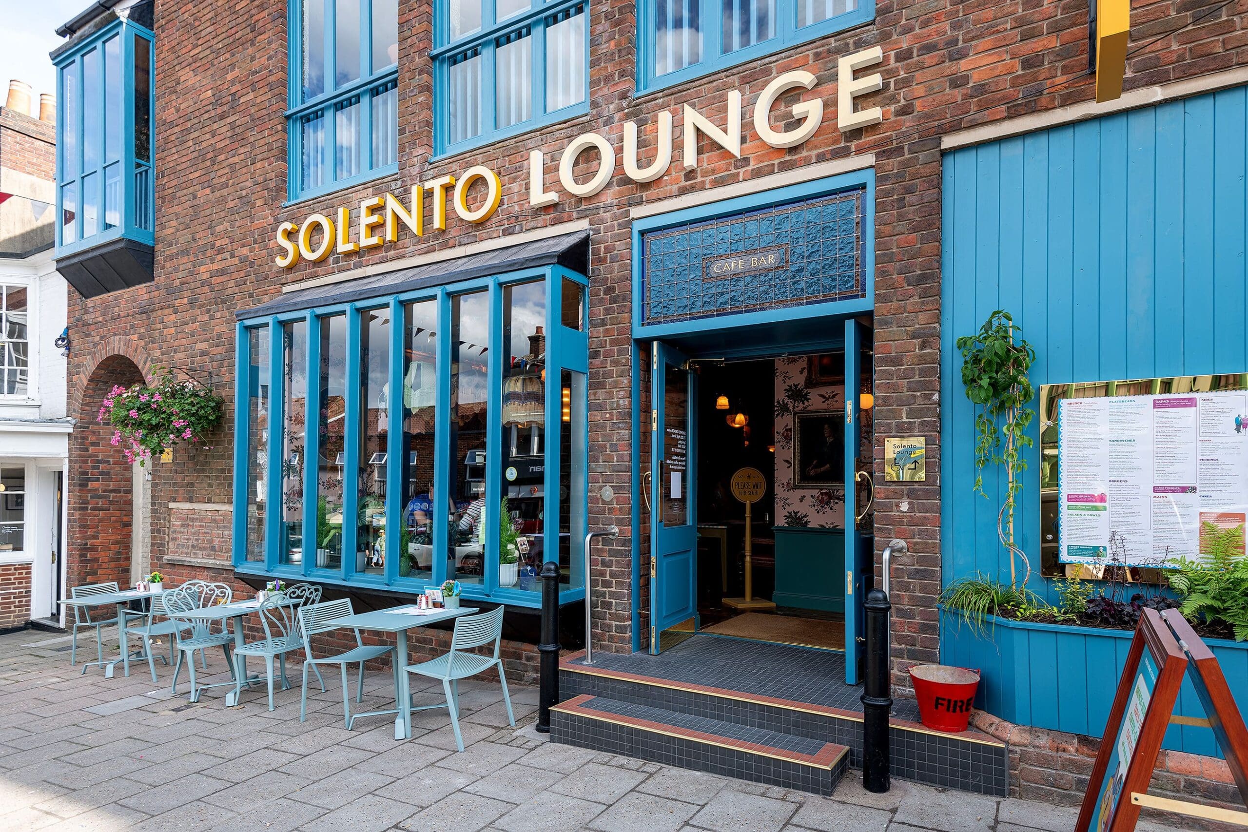 The entrance of Solento Lounge, a cozy café with large blue-framed windows, outdoor tables and chairs, a signature blue door, hanging plants, and the Solento menu displayed on the right wall.