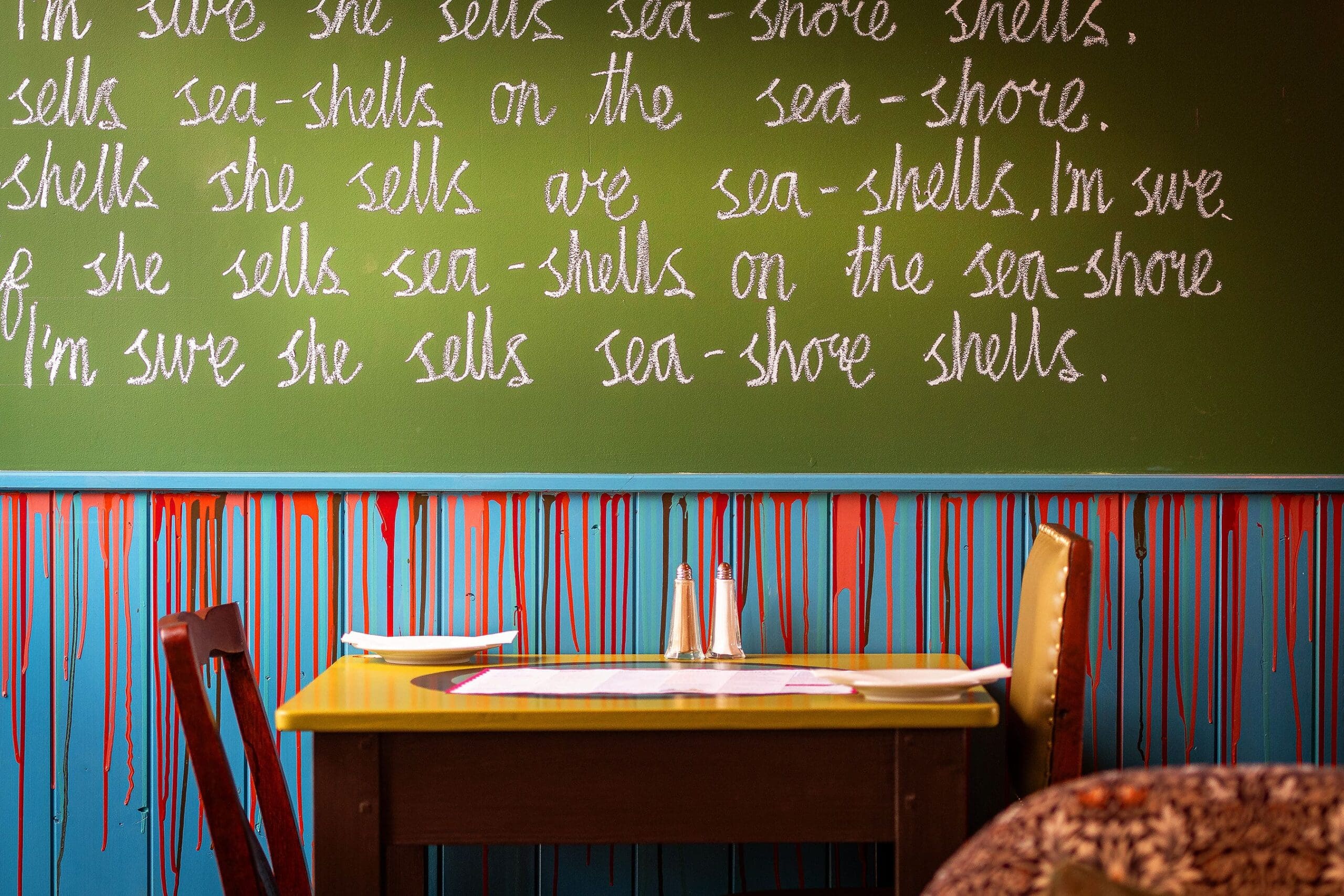 A table with two chairs sits against a wall; above it, a green chalkboard displays repeated lines of the tongue-twister "She sells sea-shells on the sea-shore." Red, blue, and Solento-yellow paint drips down the lower part of the wall.