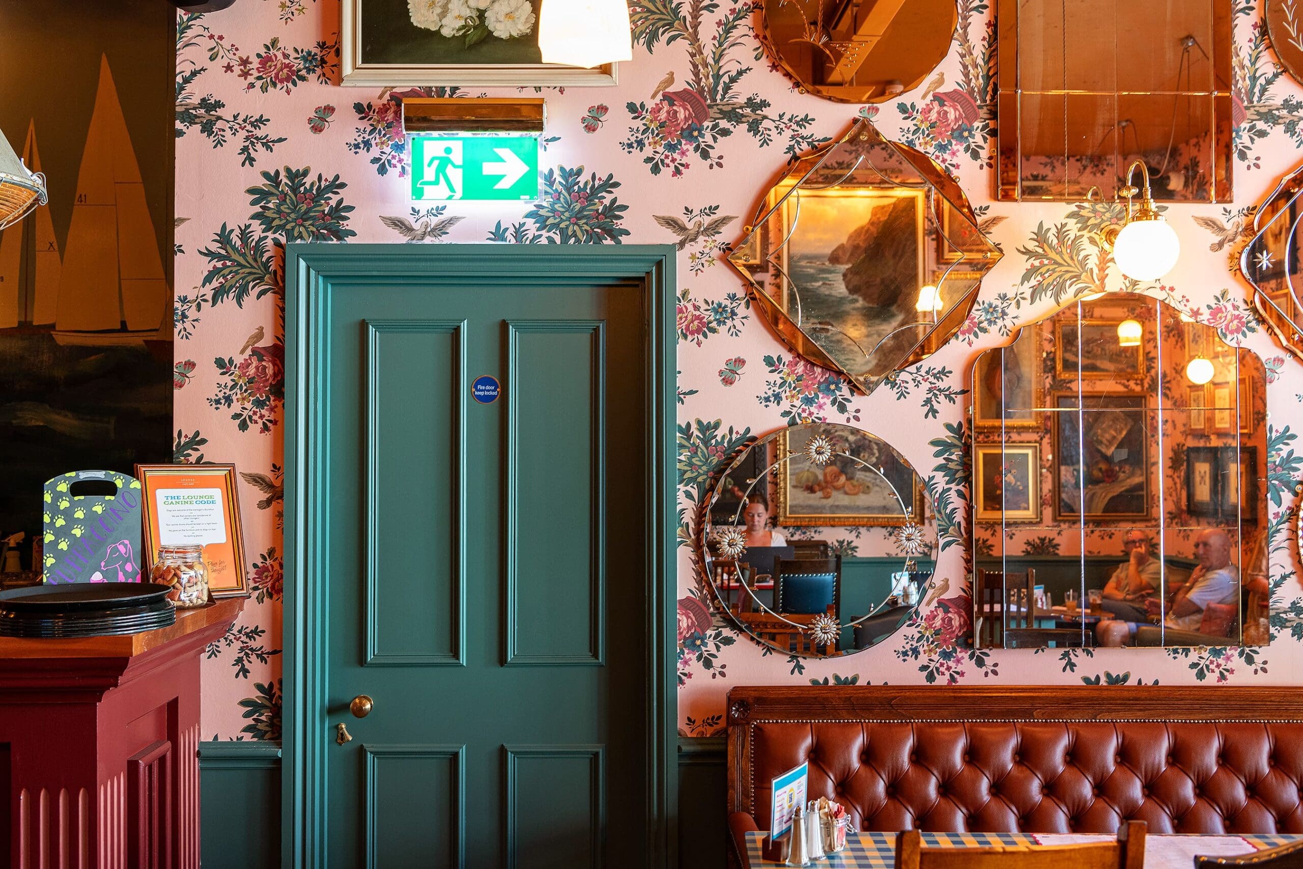 A cozy Solento room with floral wallpaper, green door, and brown leather seating. Several mirrors and paintings hang on the wall, reflecting warm lights and interior details. An exit sign glows above the door.