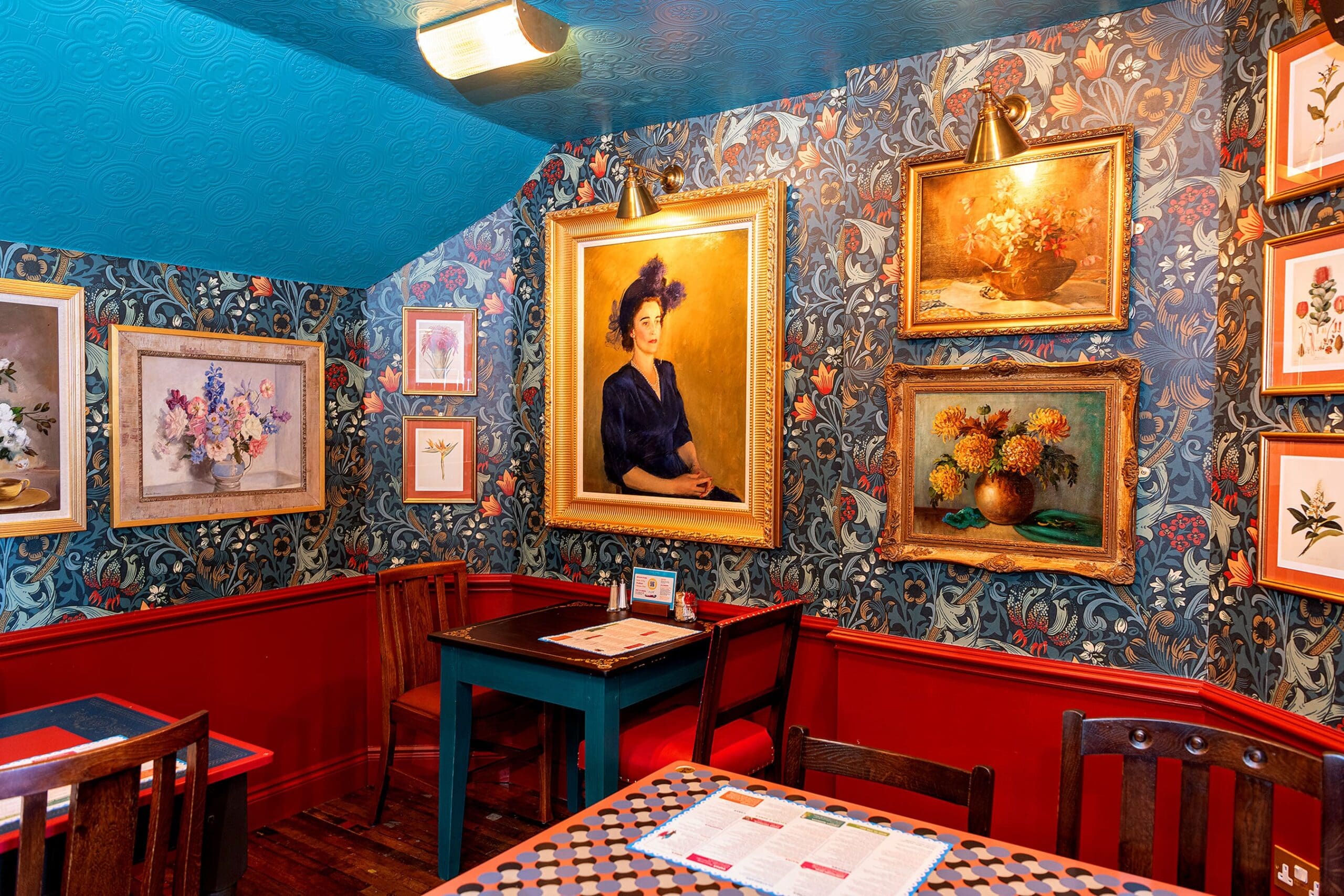 Eclectic dining room with colorful floral wallpaper, red wainscoting, Solento wooden tables and chairs, and various framed paintings, including floral still lifes and a portrait of a woman in a dark dress and hat.