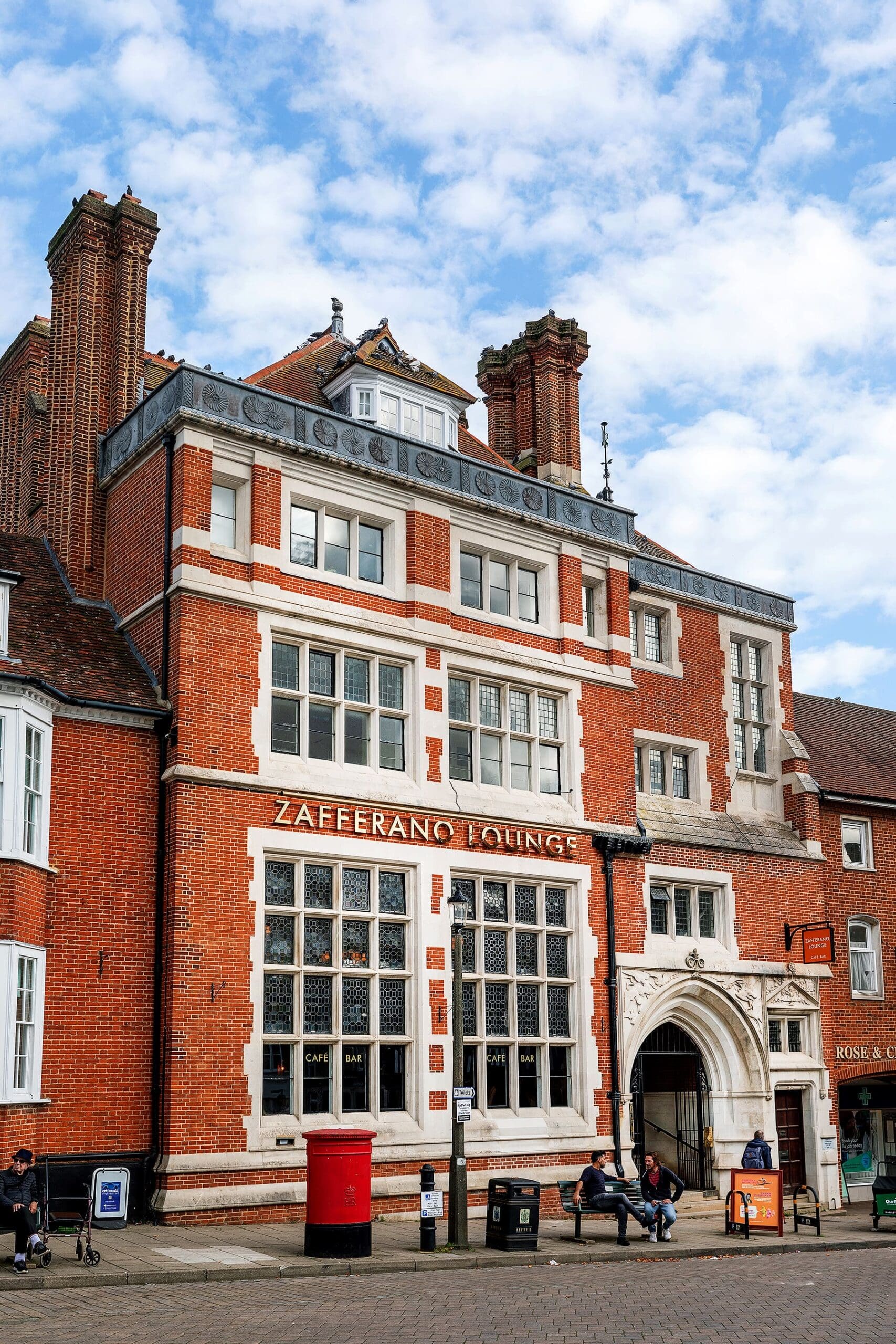 A large red-brick building with white window frames and ornate chimneys is home to Zafferano Lounge, a welcoming cafe-bar. People sit and walk outside, while a red postbox stands near the entrance of Zafferano. The sky above is partly cloudy.