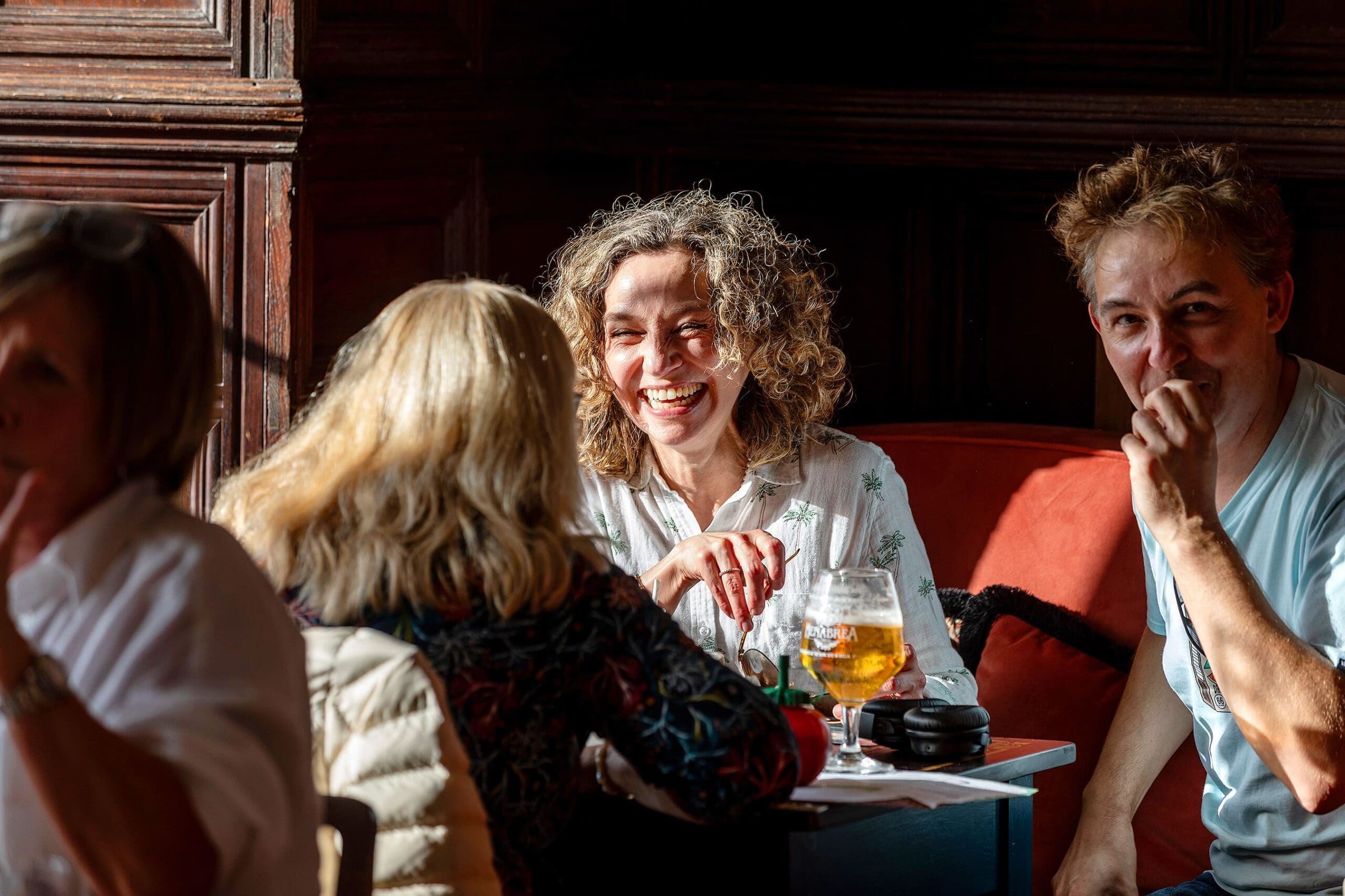 Three people sit at a sunlit restaurant table, smiling and laughing together. The woman in the center holds a zafferano-infused drink, while her friends nearby enjoy the lively conversation.