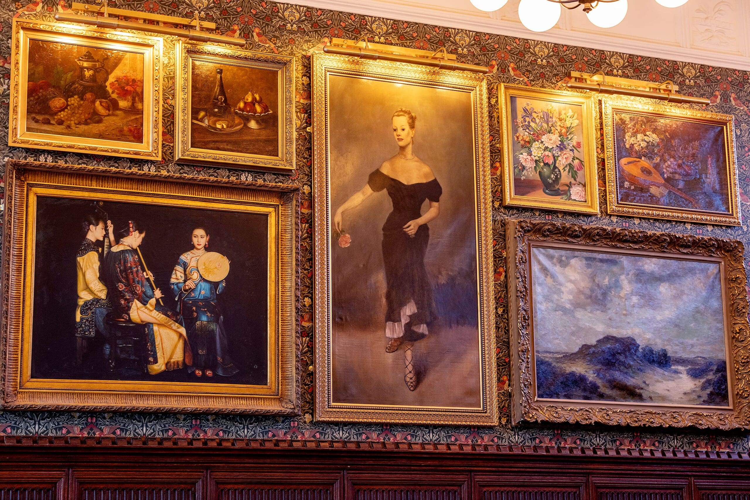 A wall adorned with ornate, gold-framed paintings—including portraits, a woman in a black dress, floral arrangements, and a zafferano-hued landscape—set above wood paneling and illuminated by warm lights.