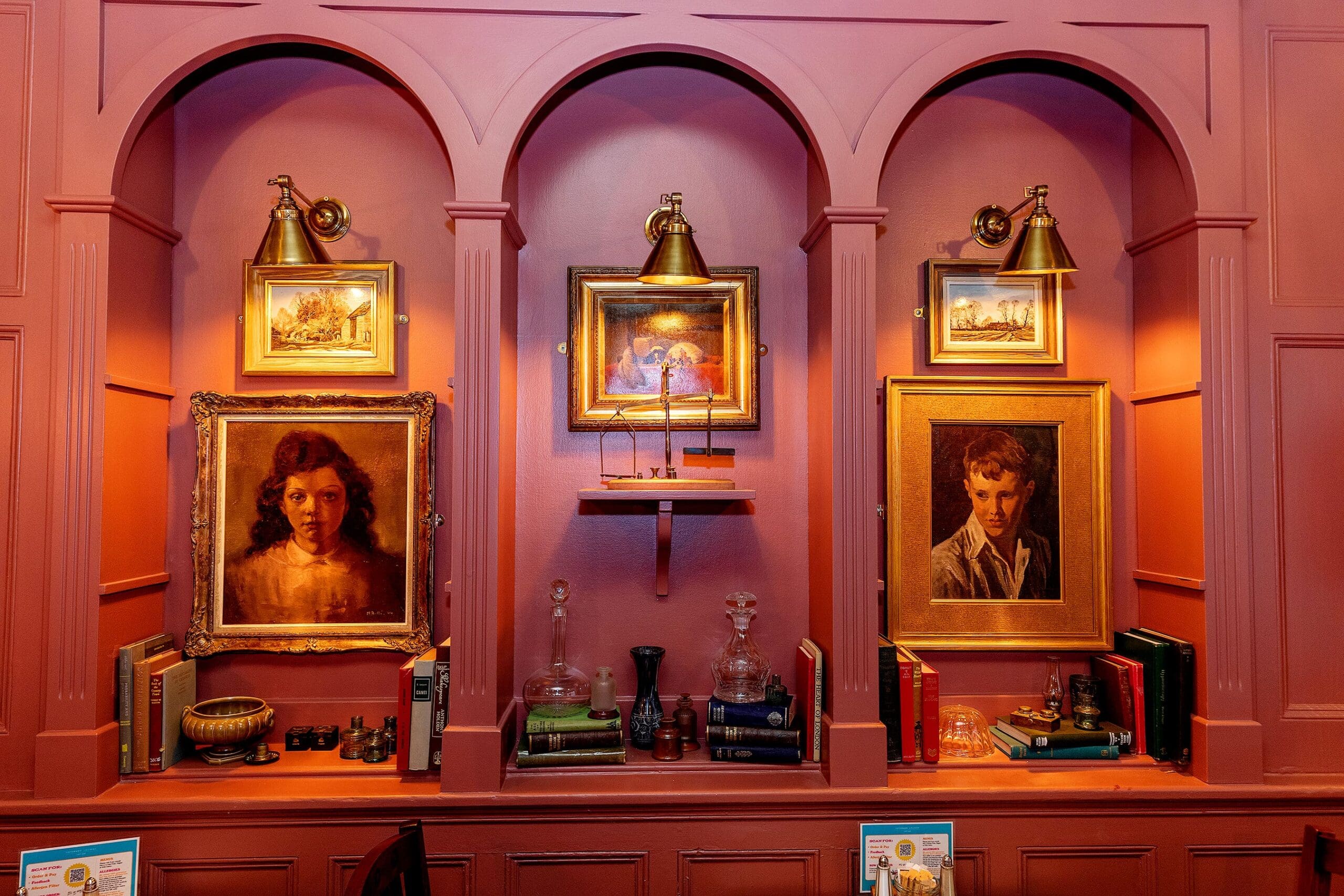 Three alcoves in a pink wall display framed portraits, zafferano-hued antique glassware, books, and small art pieces, each alcove lit by a brass wall light above the artwork.
