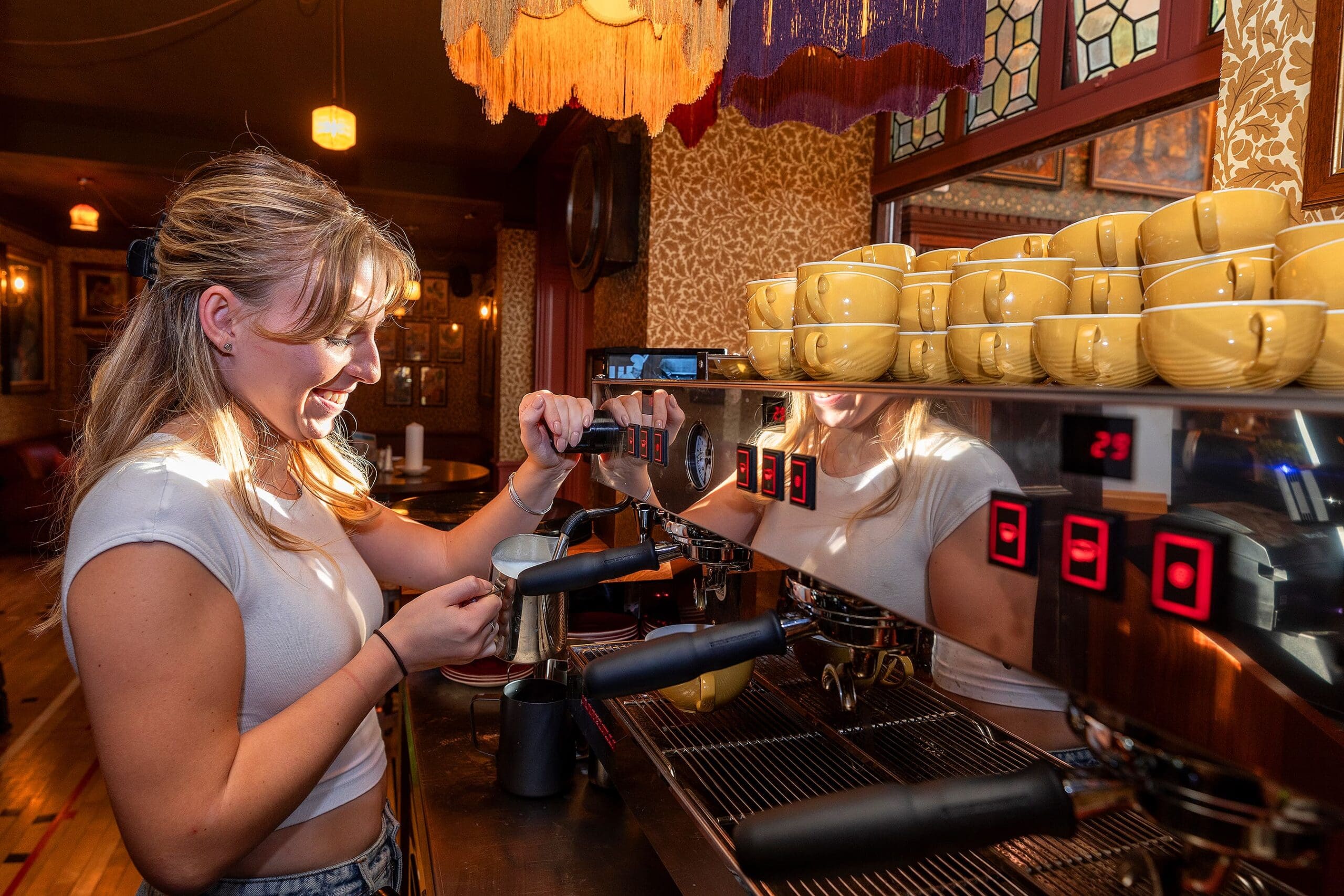 A smiling woman steams milk with an espresso machine in a cozy, warmly lit café filled with zafferano yellow cups stacked on top of the machine.