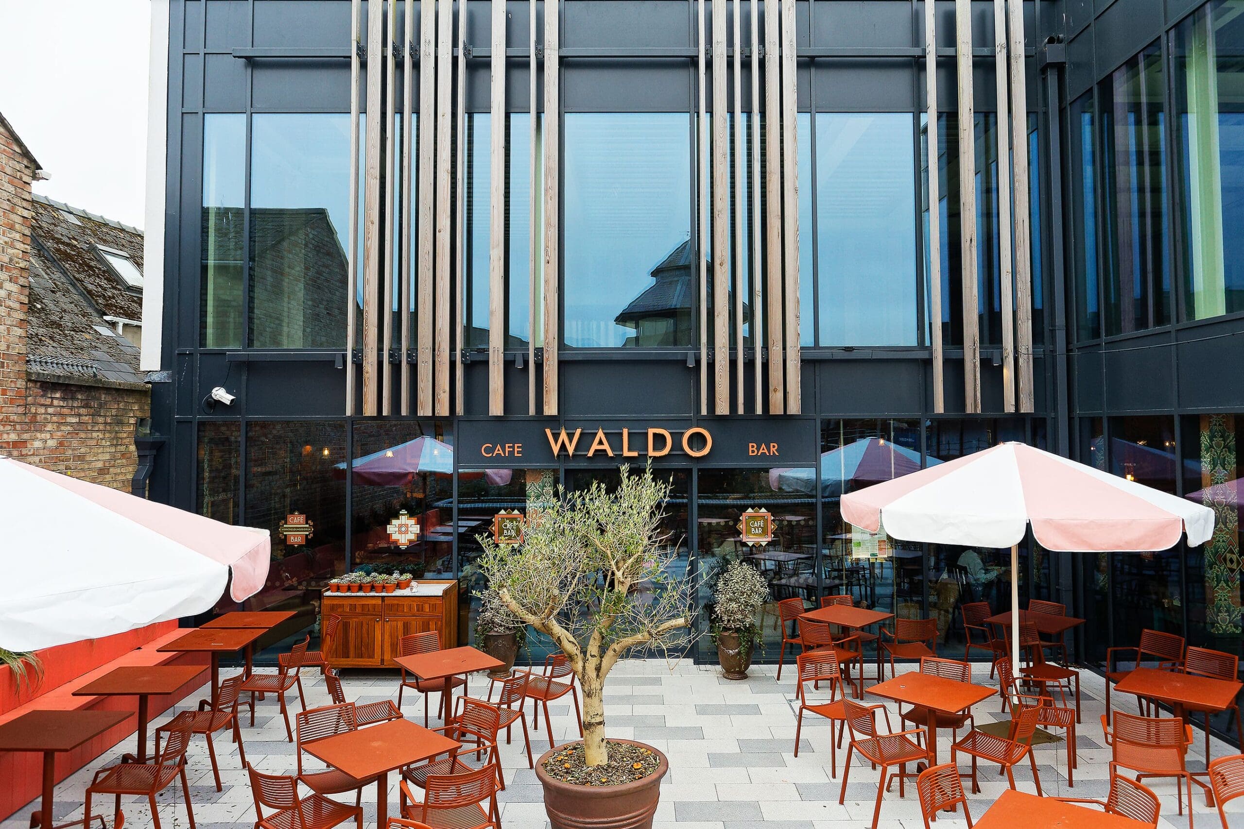 The outdoor seating area at Waldo café-bar features red tables and chairs, potted plants, and large umbrellas. The building’s striking design includes glass walls and vertical wooden slats on the upper floors.