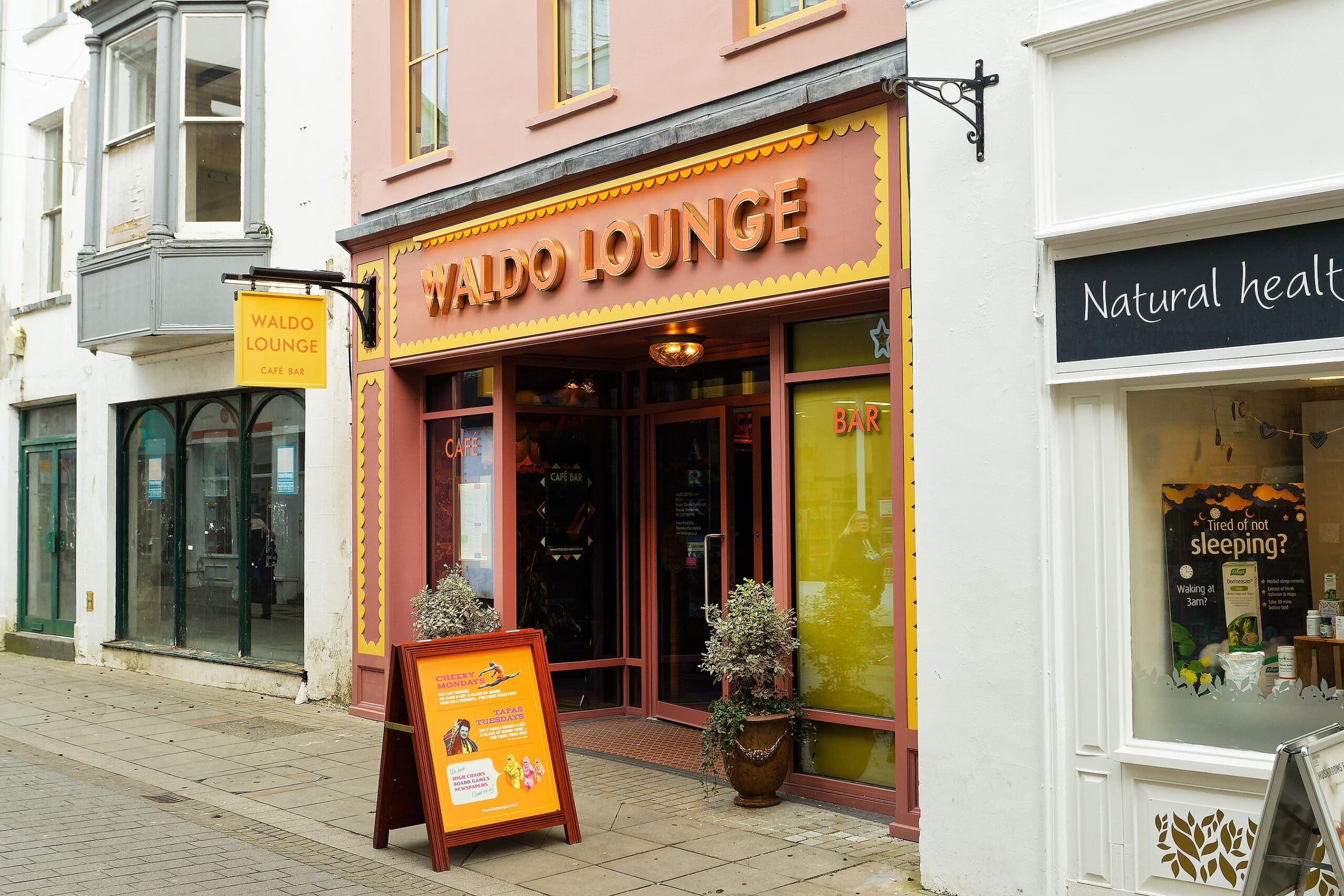 Exterior of Waldo Lounge café bar, with a bright orange and yellow Waldo sign, decorative trim, two potted plants by the entrance, and a sandwich board on the sidewalk; neighboring shops are visible.