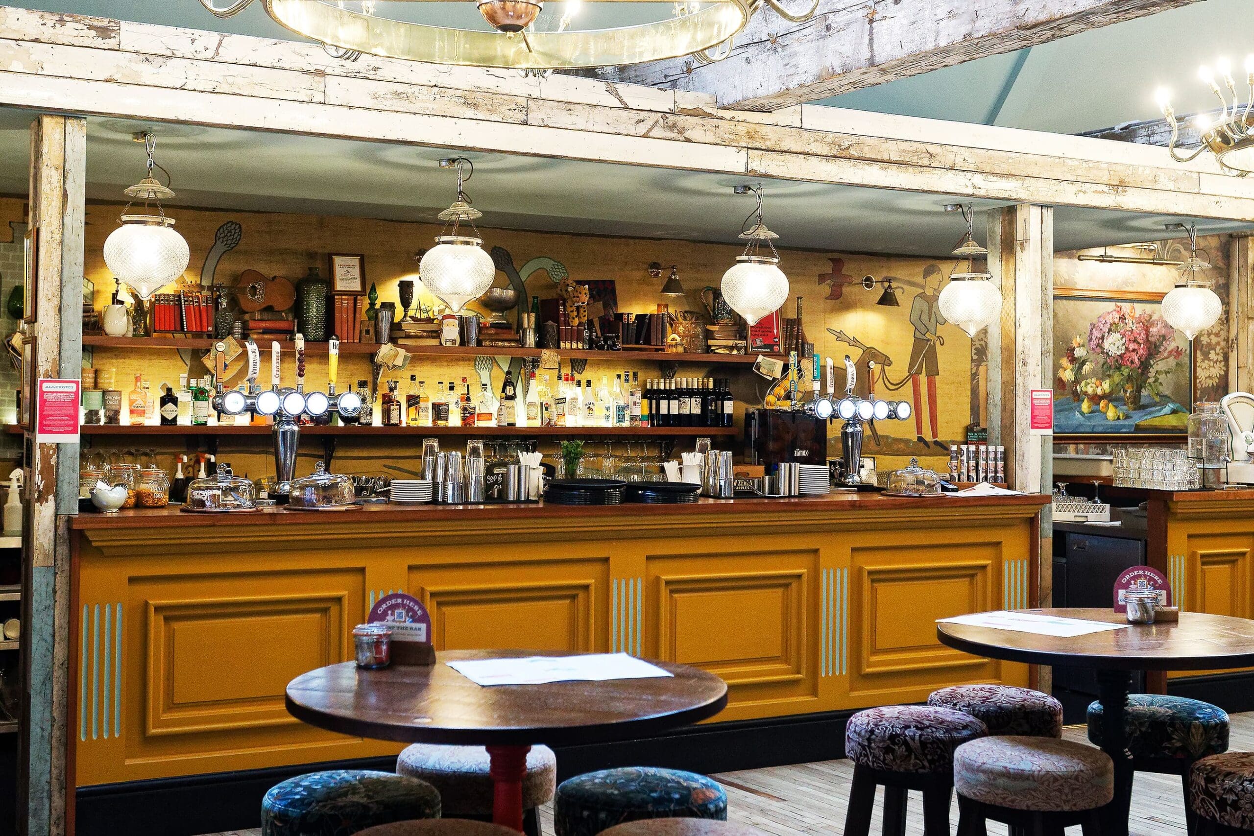 A cozy pub with a mustard yellow bar, wooden stools, hanging globe lights, and shelves filled with bottles and glasses. Eclectic art and vintage touches add warmth—and if you look closely, you might just spot Waldo hiding among the décor.