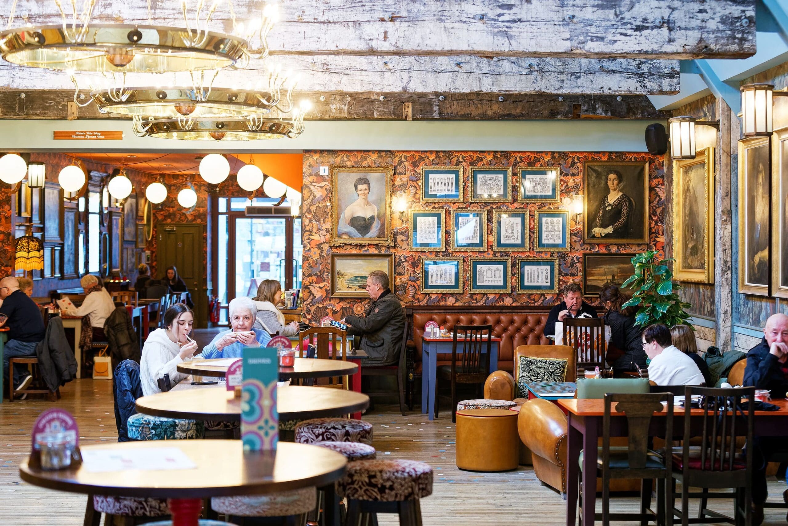 A cozy pub interior with wooden beams, chandeliers, and framed portraits on the wall—even Waldo might blend in here. Several people sit at tables and on leather couches under warm lighting, engaged in conversation or eating.