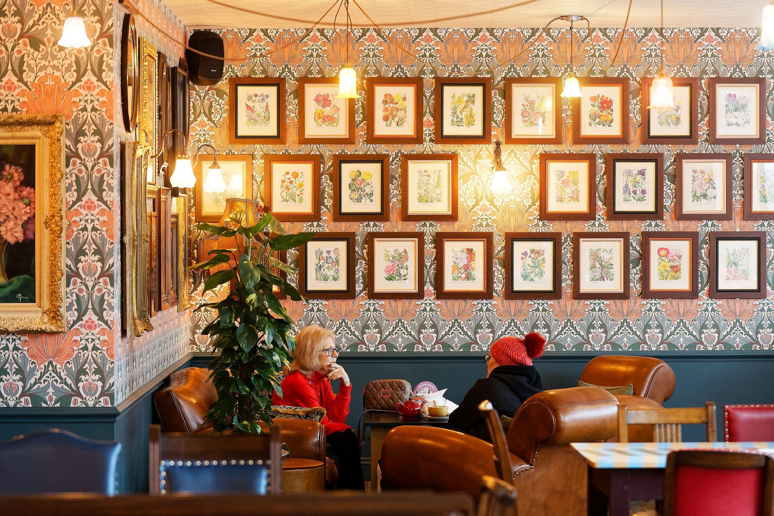 Two people sit and talk at a cozy café with leather chairs and floral wallpaper, surrounded by framed flower artwork and warm hanging lights. A tall potted plant—and a small figurine of Waldo—are next to their table.