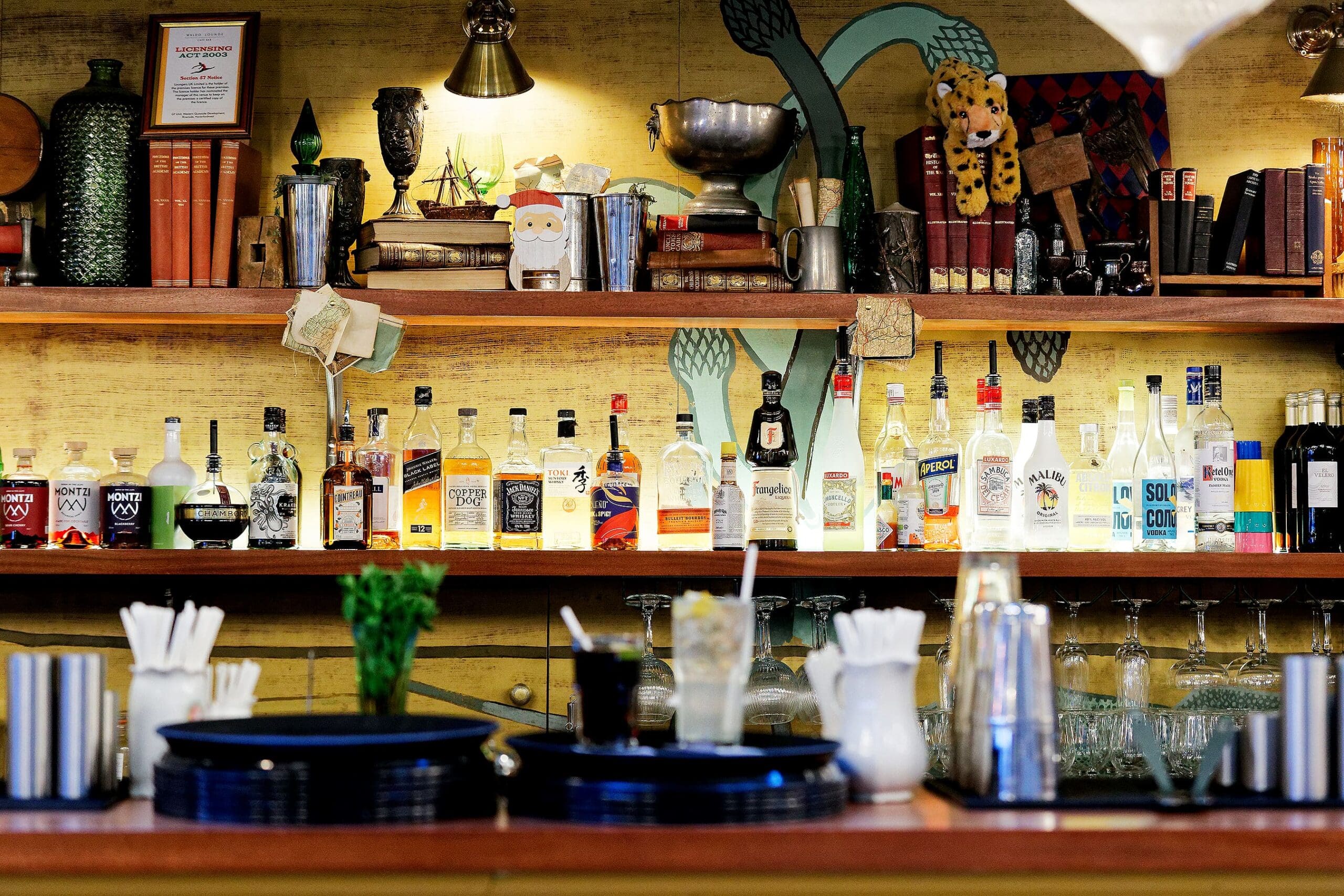 A bar counter with plates, straws, and glassware in the foreground; bottles of various alcohols and books—where you might even spot Waldo—are neatly arranged on wooden shelves, with eclectic decor and ornaments in the background.