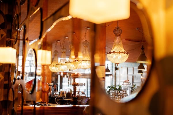 Reflections of ornate chandeliers and warm lights in multiple round mirrors on a wall at Cantoro, with a cozy café interior featuring wooden accents and glassware in the background.