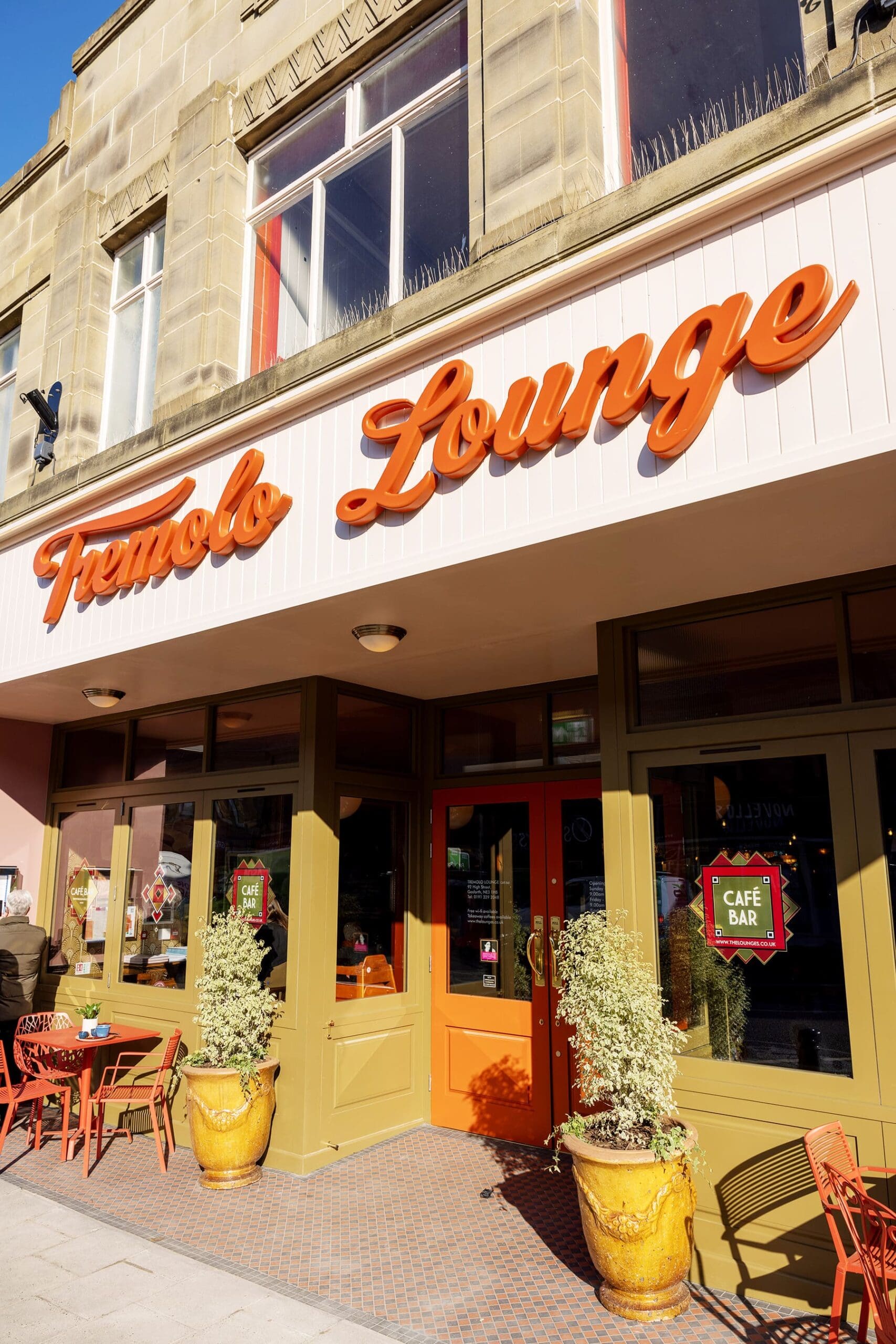 Cafe exterior with an orange sign reading "Tremolo Lounge," large windows, tremolo-orange framed doors, two potted plants, and outdoor seating with matching orange chairs and tables on a sunny day.