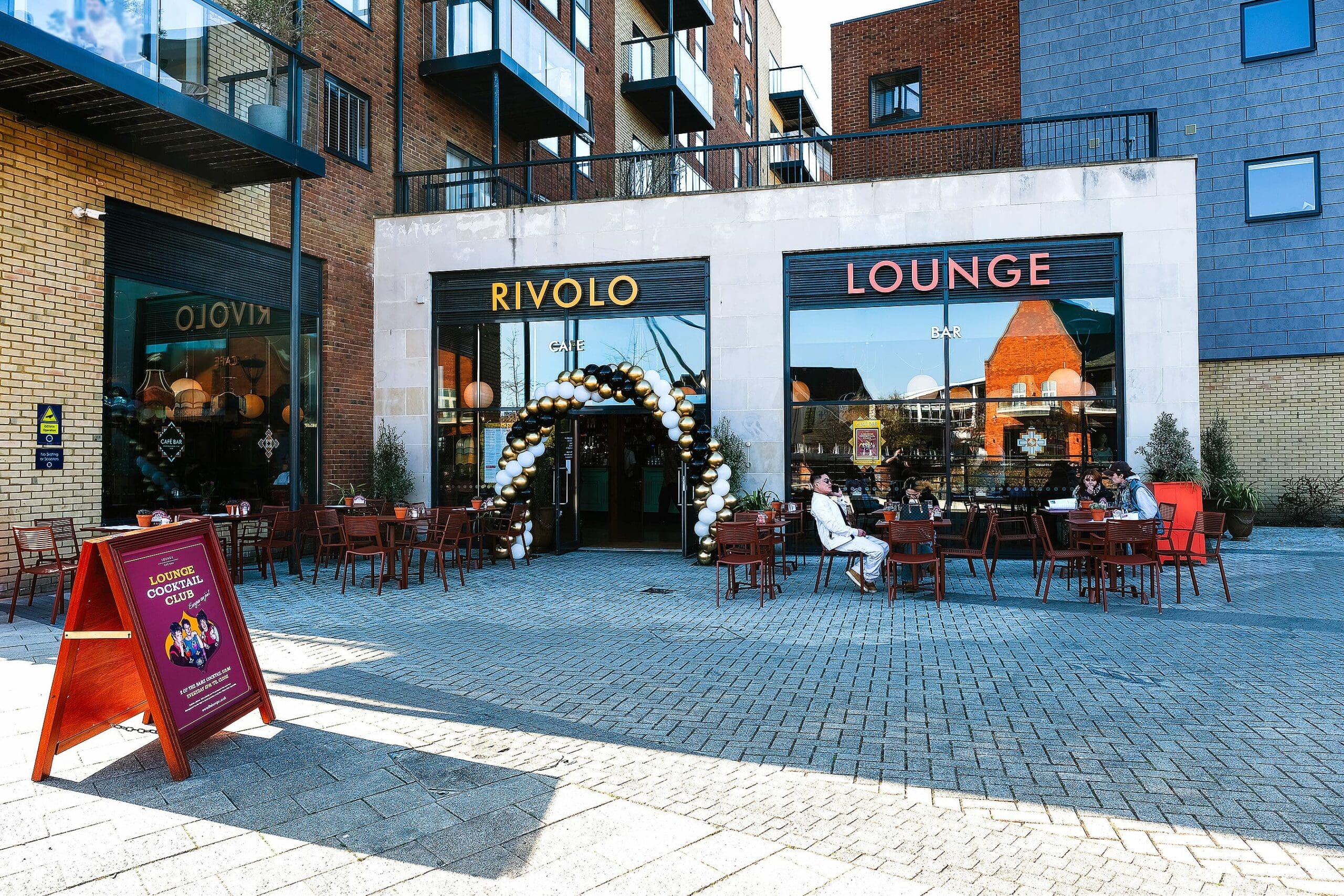 Outdoor seating area of Rivolo Lounge with people dining at tables. The entrance, adorned with a black, white, and gold balloon arch, welcomes guests to Rivolo’s grand opening. Modern apartment buildings rise in the background.