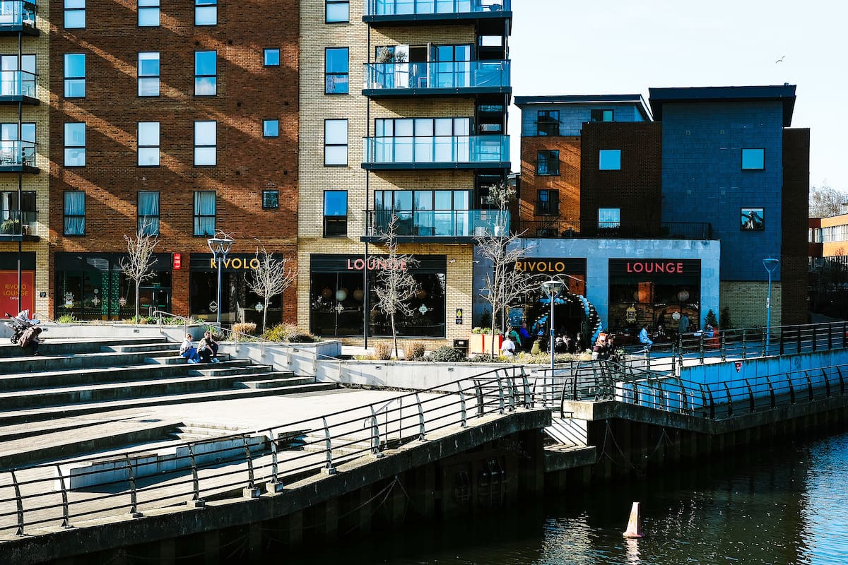Modern riverside apartment buildings with balconies and ground-floor cafes overlook the rivolo. Steps and railings lead down to the water, where a few people are relaxing in the sunlight. Leafless trees line the walkway.