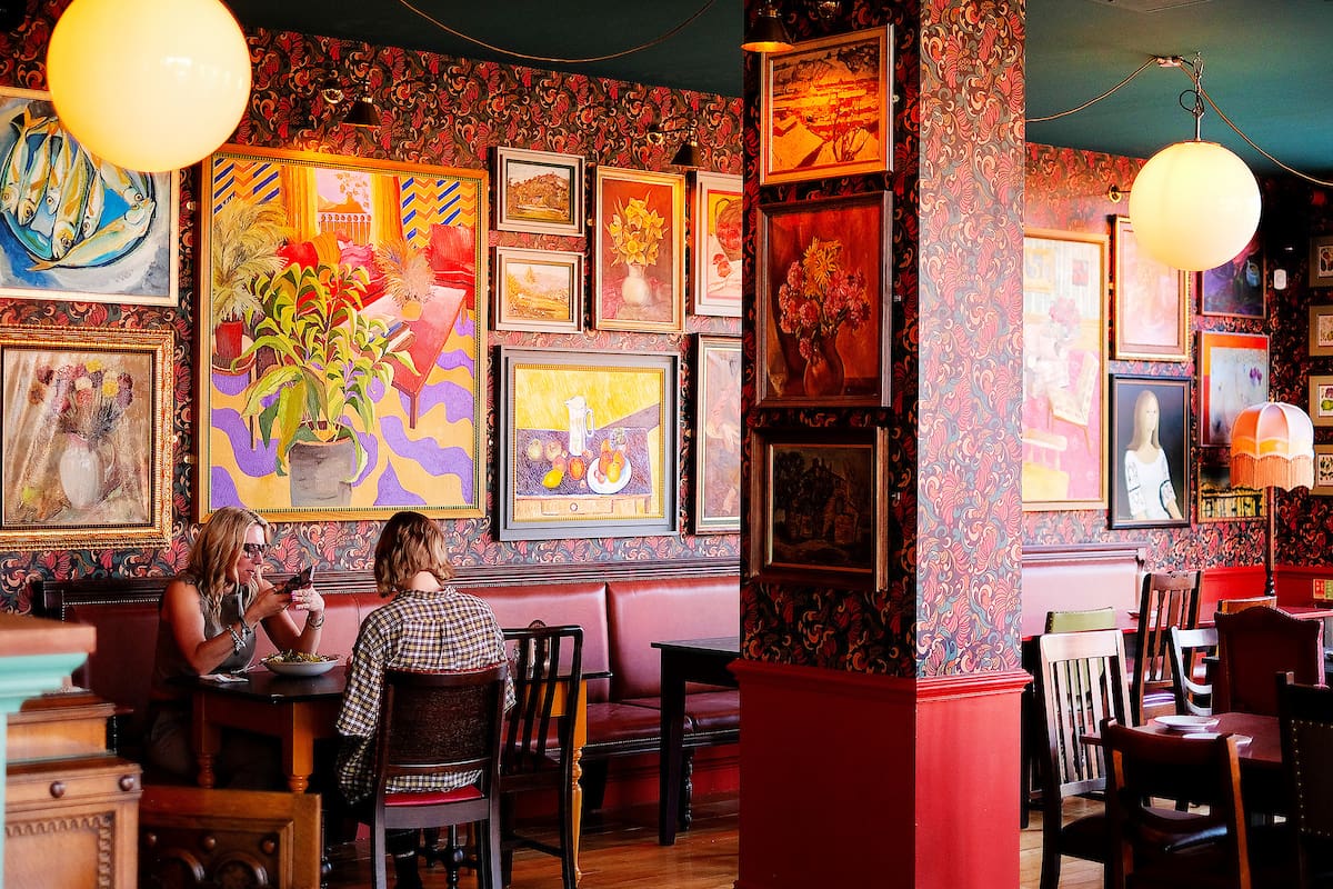 Two women sit at a table in a cozy, art-filled café with patterned wallpaper, red accents, and vibrant rivolo paintings covering the walls. Wooden chairs and empty tables fill the inviting, eclectic space.