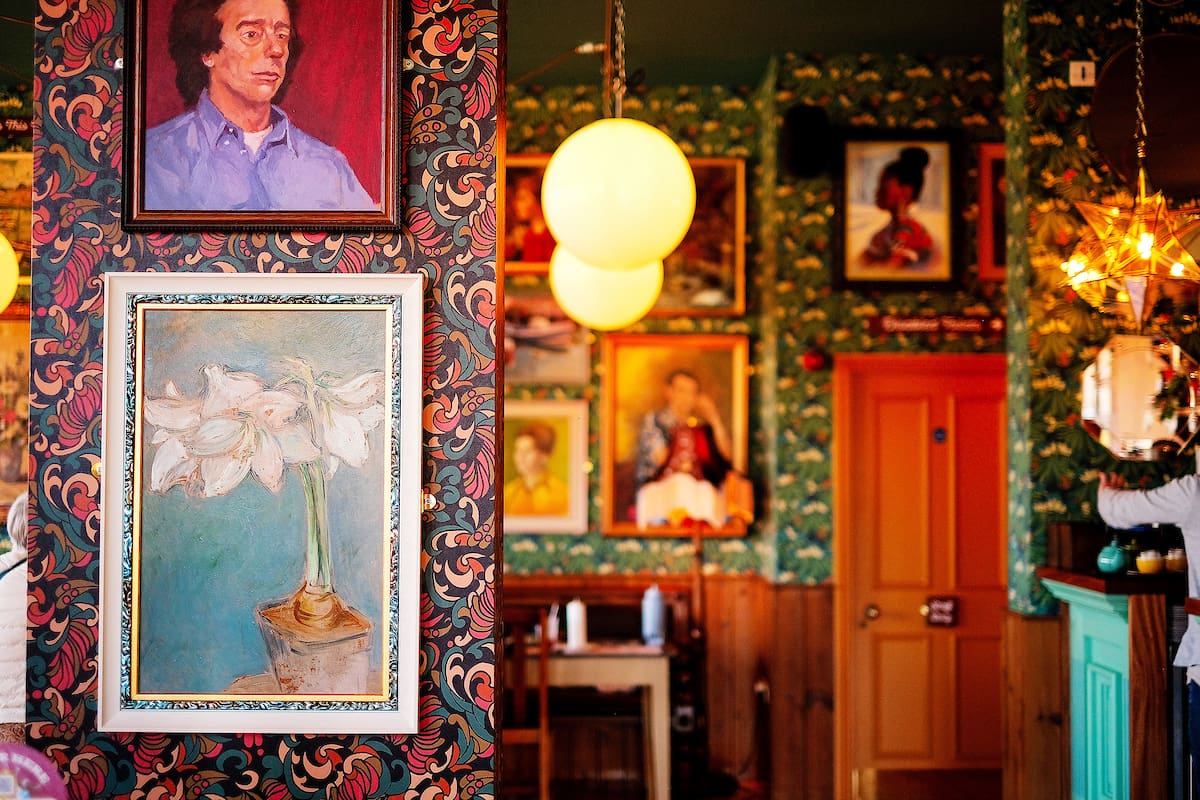 Colorful interior of a room with rivolo-patterned wallpaper, framed paintings on the walls, two round hanging lights, and a wooden door; a hand is seen on the right side near a counter.