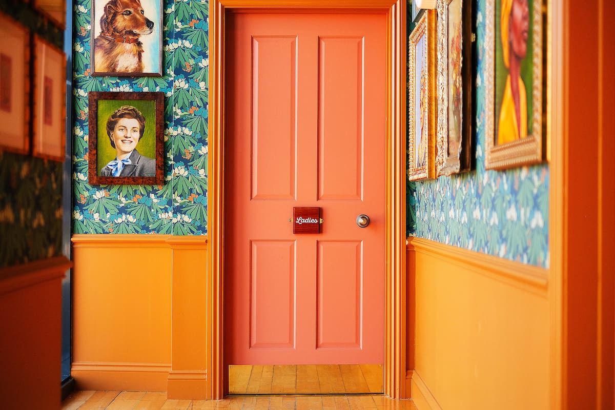 A coral-colored door with a small red rivolo sign is set in a hallway with floral wallpaper and orange trim. Framed portraits of people and a dog decorate the walls, above the light wood floor.